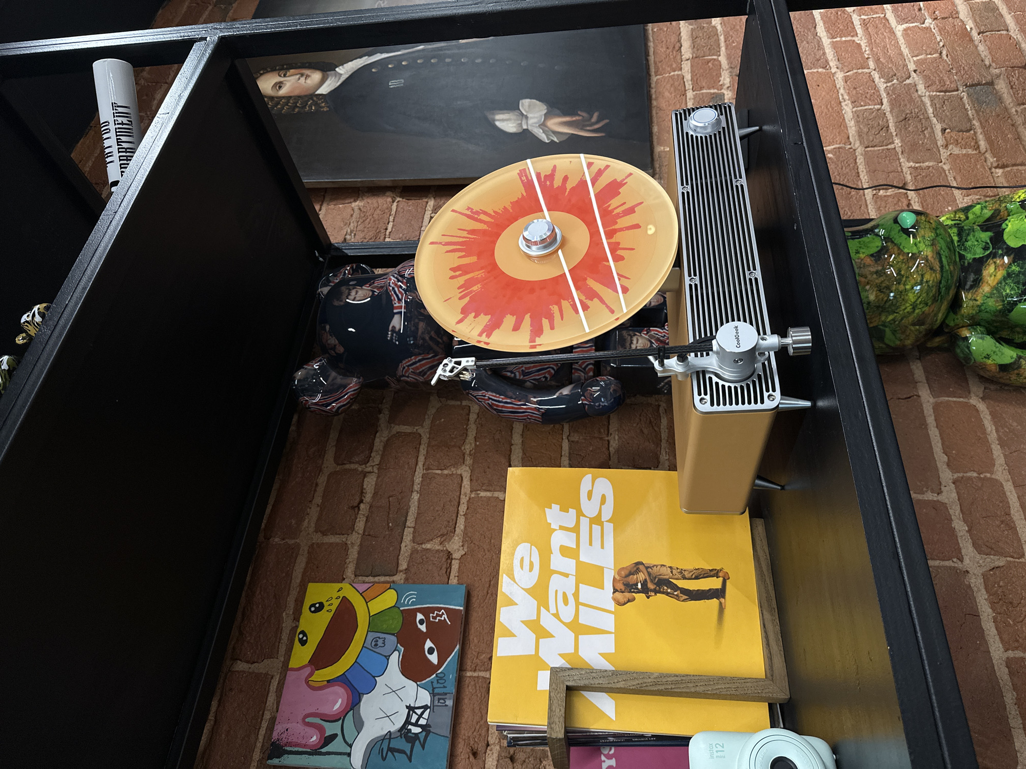 The photograph depicts a shelf unit filled with various objects, captured from a high-angle perspective. The shelf unit is constructed of dark-colored material with visible brickwork in the background, suggesting an interior space.
On the top shelf, there is a black-framed picture of a portrait, a patterned object resembling a large, reclining figure, a turntable with a yellow record, and a small, green, frog-shaped sculpture.
The shelf below holds a stack of items, including a yellow album cover with the text "We Want MILES" prominently displayed. A colorful art piece is adjacent to the album. A white instant camera is also visible at the bottom shelf.