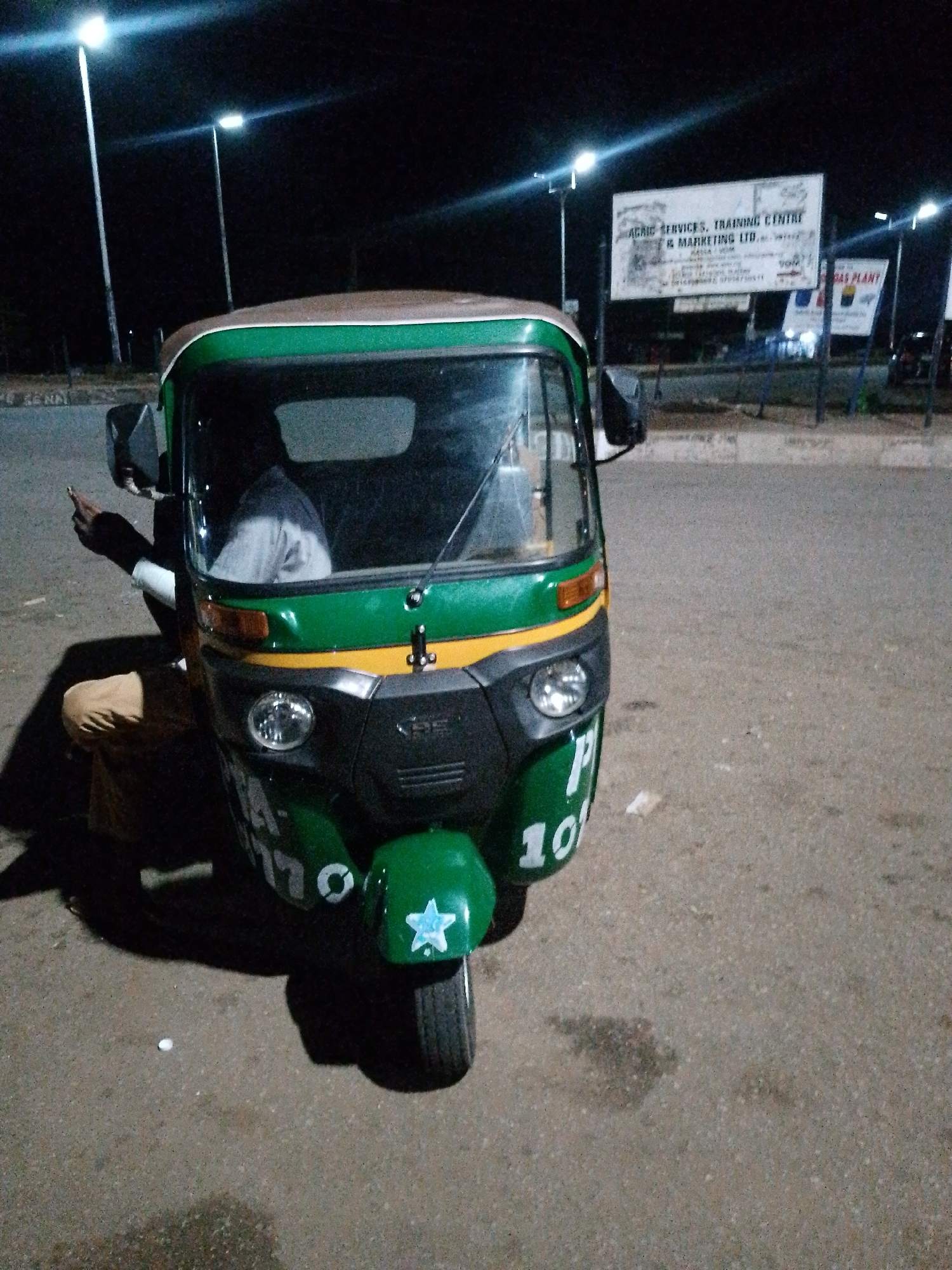 This media file captures an auto-rickshaw parked on a paved surface, likely a road or parking lot, in Bukuru, Nigeria, during nighttime. The scene is illuminated by streetlights, casting a dim glow and creating long shadows. The auto-rickshaw is predominantly green with yellow and brown accents. A person, partially visible, is seated in the driver's seat, wearing a grey and white shirt. Their left arm is extended outwards, holding what appears to be a cigarette. Another person, wearing brown trousers, is seated on the ground to the left of the auto-rickshaw, their posture suggesting they are waiting or resting.
In the background, several billboards are visible. One large billboard displays text that includes "AGRIC SERVICES, TRAINING CENTRE & MARKETING LTD." and appears to have other information below it, though it's difficult to read clearly. Another billboard to the right has text that includes "GAS PLANT". The streetlights are tall and modern, suggesting a developed area. The ground is paved and shows some signs of wear and possibly a small puddle or stain. A small white object, possibly a piece of debris or trash, is visible on the ground in front of the auto-rickshaw. A white star shape is also visible on the front of the auto-rickshaw. The overall impression is that of a quiet, somewhat industrial or commercial area at night, with the auto-rickshaw and its occupants being the primary focus. The dim lighting and the posture of the people suggest a moment of pause or waiting.