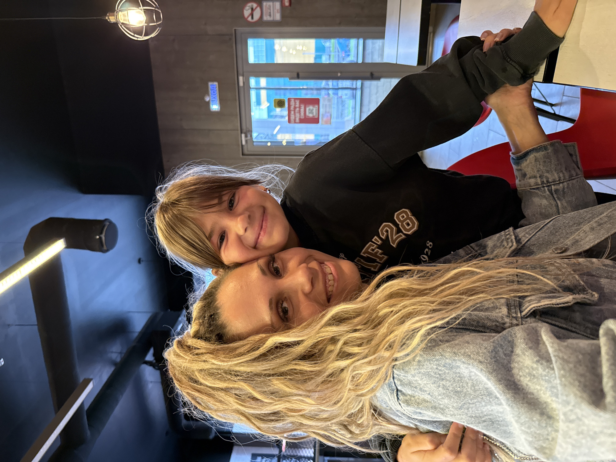 The image shows two individuals, an adult woman and a child, posing for a photograph indoors, likely in a public establishment. The woman, on the left, has long, blonde, wavy hair pulled into a high ponytail and is wearing a light-wash denim jacket. She is smiling broadly, with her head tilted slightly. The child, on the right, has shoulder-length brown hair and is wearing a dark sweatshirt with a light-colored graphic design on the front. The child is also smiling and has an arm around the woman's shoulders.
The background suggests an indoor setting with industrial-style lighting fixtures on the ceiling and dark, possibly concrete walls. There are glass doors and windows visible, with some signage. The overall atmosphere appears casual and possibly a restaurant or a similar public space. There is no specific visual information in the image to confirm the location as Ignatovo, Russia.