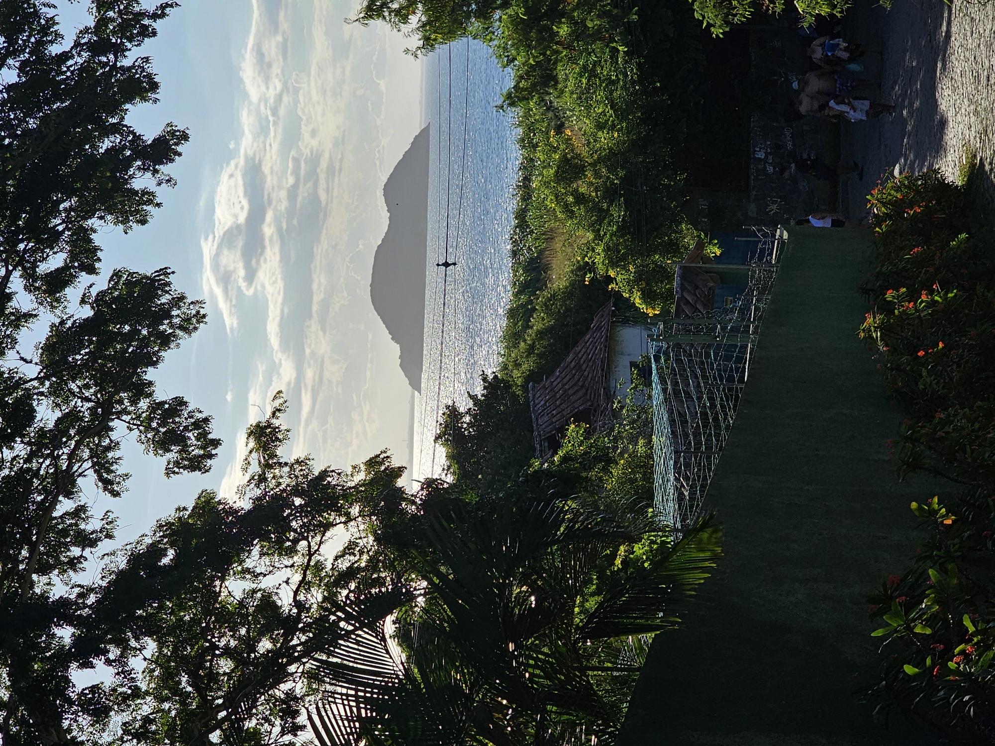 The image captures a coastal scene in Armação dos Búzios, Brazil, framed by lush foliage. A shimmering body of water stretches towards a mountainous silhouette on the horizon, partially obscured by clouds. Power lines run horizontally across the water. A structure, possibly a house, is visible amidst the greenery. On the right, people are walking on a paved pathway. The scene is bathed in sunlight.