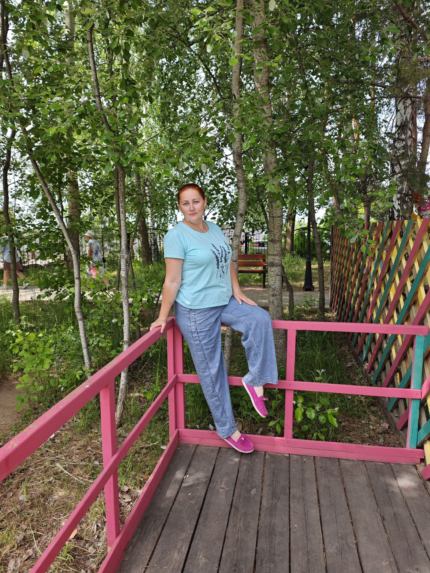 The image depicts a woman with red hair, wearing a light blue t-shirt and jeans, sitting on the railing of a wooden structure. She is also wearing pink shoes. The structure appears to be a small deck or platform, painted pink. Behind her are trees and a colorful fence. The background suggests a park or wooded area. The image was taken in the city of Chaikovsky, Russia. Two other people appear to be in the background walking by a path. There is a bench behind the woman and a black light post in the same direction. The ground is covered in woodchips.