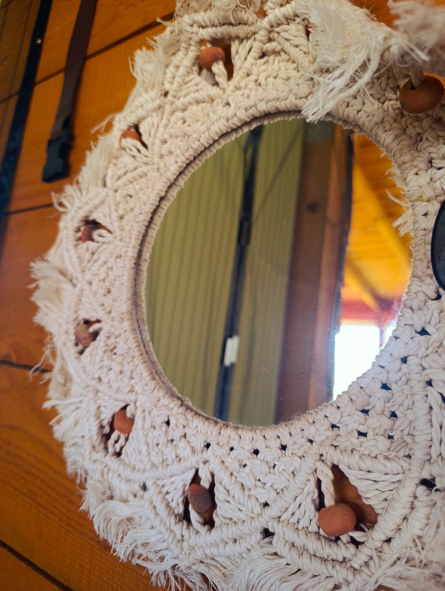 The image shows a round mirror with a macrame frame decorated with wooden beads. The mirror is reflecting parts of a room with wood paneling and a bright yellow ceiling. In the upper left of the image, part of a strap with a buckle is visible against the wood paneling. The macrame is off-white and has a fringed edge. The wooden beads are brown. The scene does not provide any information about the location being in Shiroko pole, Bulgaria.