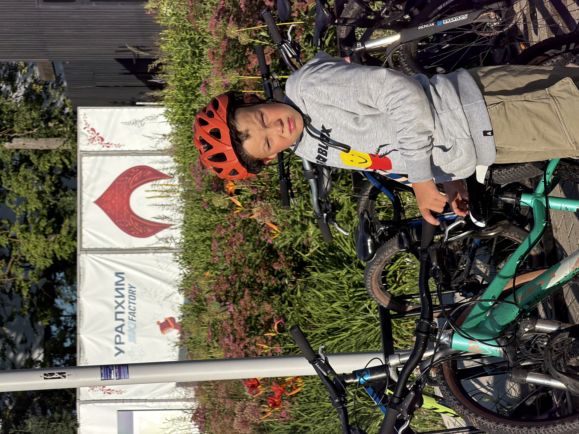A young boy wearing a red bicycle helmet and a grey sweatshirt with a cartoon character on it stands in front of several parked bicycles. The boy is looking directly at the camera with a serious expression. He is wearing tan shorts. Behind him, a row of bicycles is parked in front of a fence with a large banner that reads "УРАЛХИМ SKI FACTORY". The banner has a stylized red graphic. The scene appears to be outdoors, with green foliage and flowers visible in the background. The lighting suggests it is daytime, possibly late afternoon due to the shadows. The location is identified as Odintsovo, Russia.