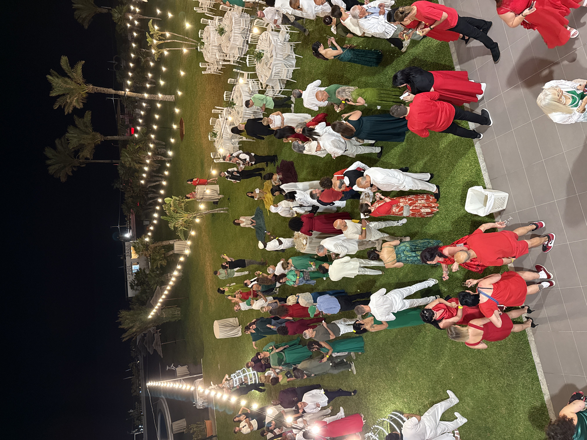 The image captures an outdoor event at night, likely in Isola delle Femmine, Italy, suggested by the presence of palm trees and possibly nearby coastal features. A large group of people is dispersed across a grassy area, some mingling near round tables adorned with white linens, indicating a possible reception or banquet setting. Many individuals are dressed in varying shades of red, green, and white clothing. Several string lights are hung above the scene, providing illumination. Some individuals appear to be in conversation, while others are more isolated in their actions. The scene transitions to a tiled or paved area on one side, adding another element to the environment.