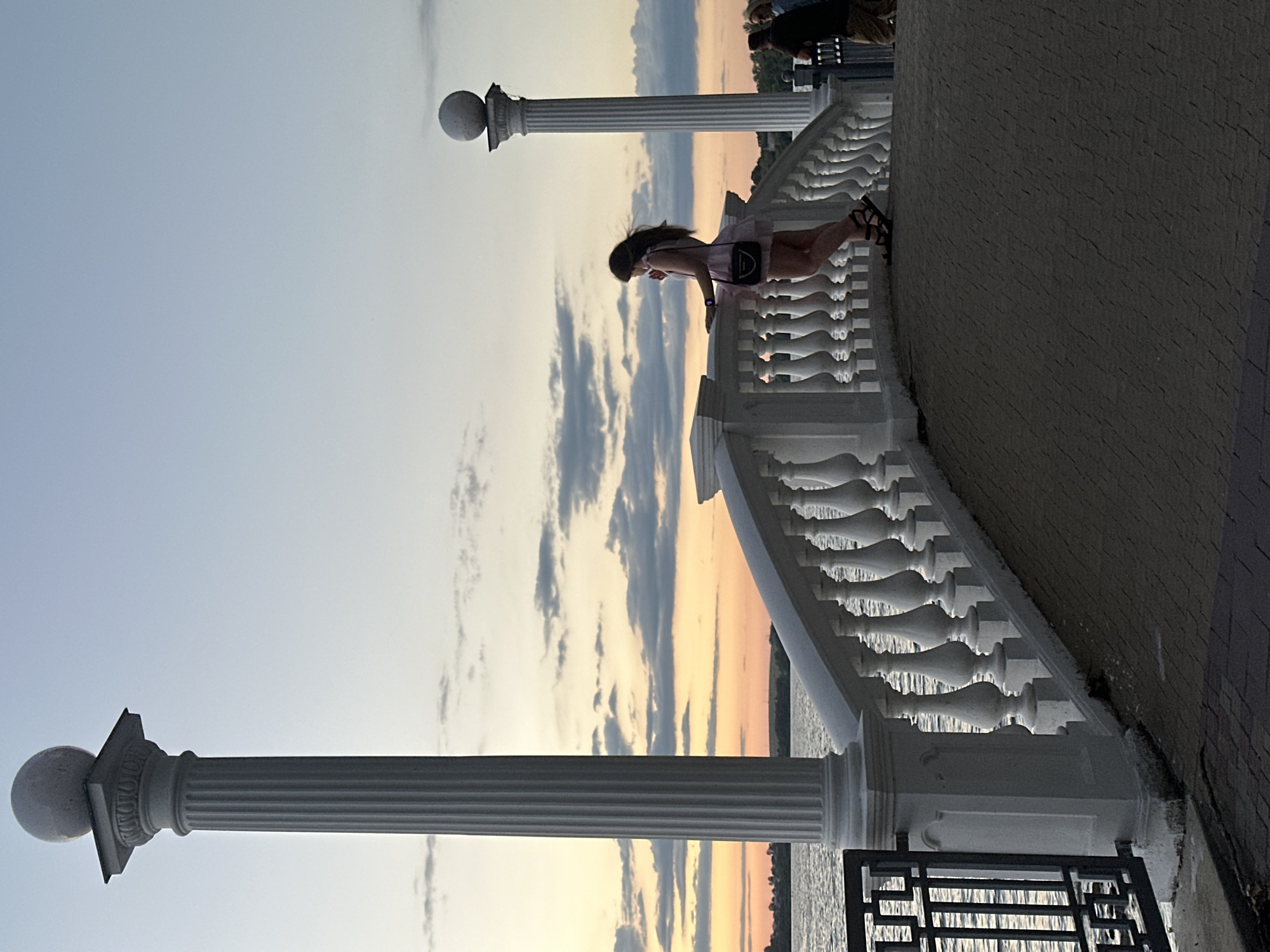 The image depicts a twilight scene on a promenade overlooking a body of water, likely Ternopil Pond, in Ternopil, Ukraine. Two prominent white classical columns with spherical finials frame the view. In the foreground, a white balustrade curves along the promenade, leading the eye towards the sunset. A young woman in a light-colored dress and black sandals stands on the promenade, facing away from the camera, gazing at the water and the colorful sky. She is holding a small black purse. Further to the right, two other individuals are partially visible, also on the promenade. The water is calm and reflects the soft hues of the sunset, which features orange and pink tones against a pale blue sky with scattered gray clouds. The ground of the promenade is paved with dark interlocking bricks. A dark metal railing is visible on the far left, separating the promenade from the water's edge.
