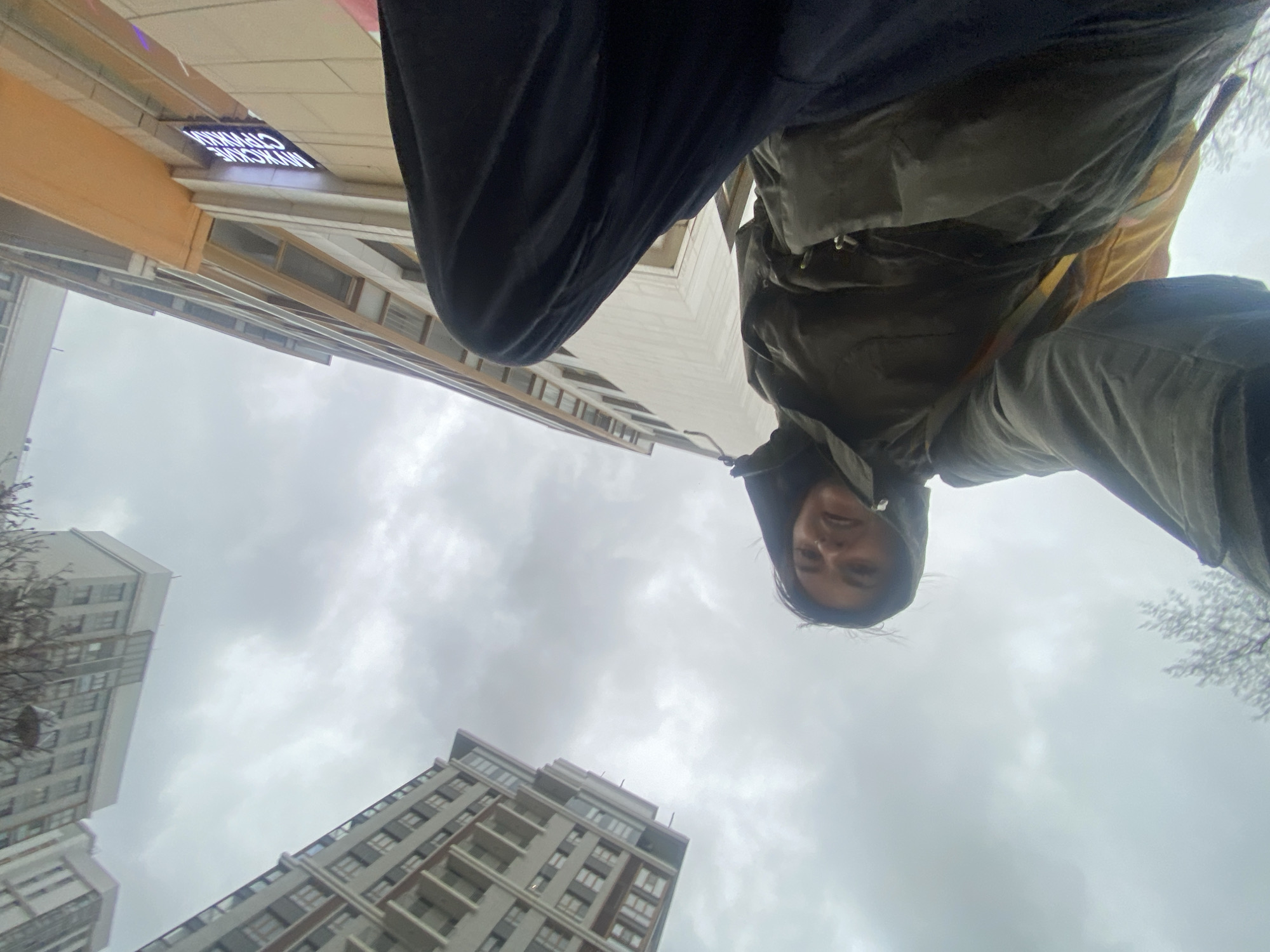 The image presents a low-angle, perspective view of a person standing in an urban environment. The buildings and sky are visible.
Specifically, a man in dark pants and a hooded jacket is positioned in the foreground. Part of a building is visible with the sign reading "MYKCARE CHATHAM". In the distance, there are several residential buildings under a cloudy sky. Based on the style of the buildings and signage, the location is Moscow, Russia.