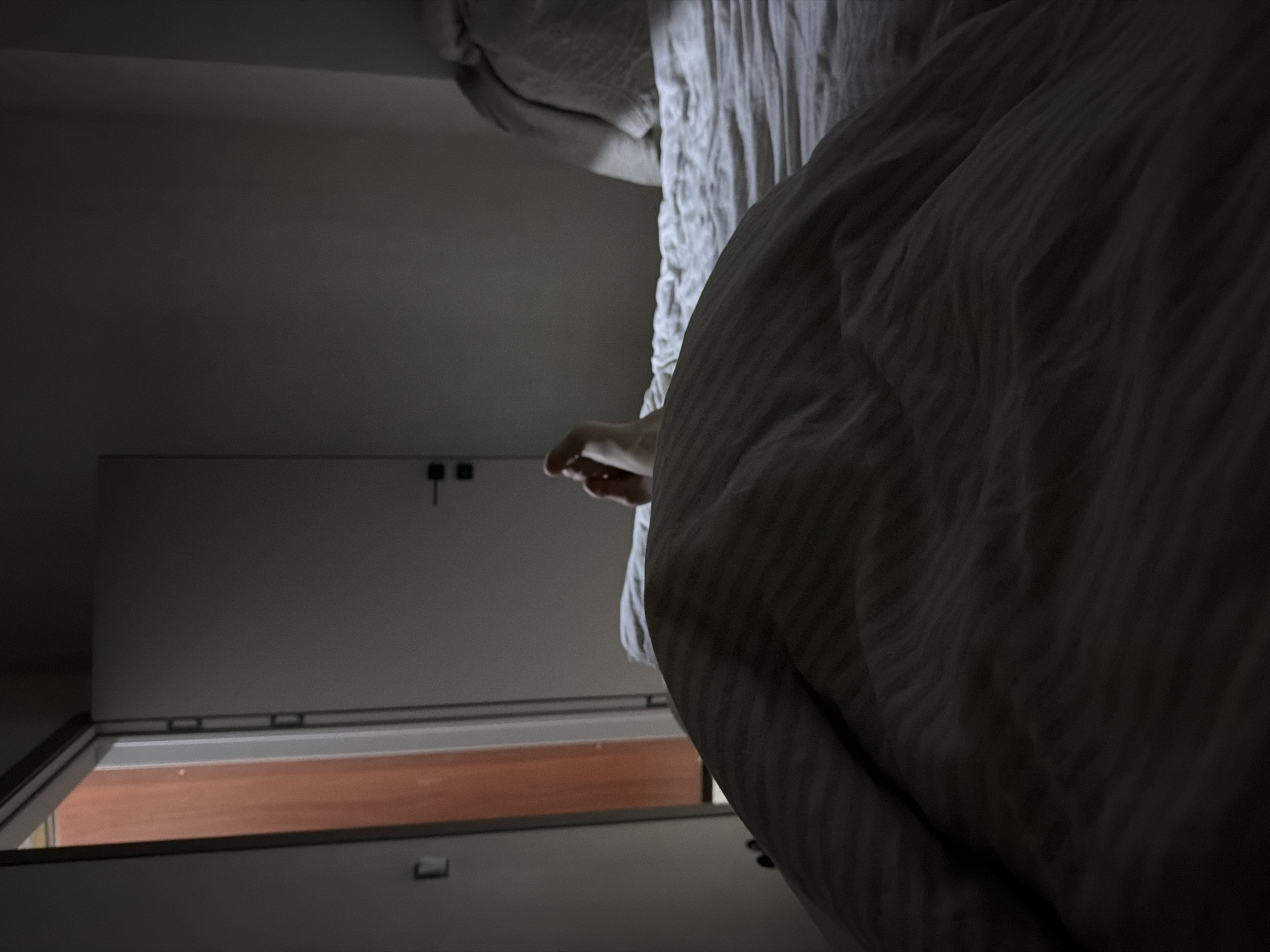 The media file captures an intimate, dimly lit indoor scene, likely a bedroom, with the camera angled from within a bed. The image is initially rotated 90 degrees clockwise, but when viewed upright, a person's hand is visible emerging from underneath a light-colored, subtly striped duvet, suggesting someone is resting or waking up in bed. The bed occupies the right side of the frame, with wrinkled sheets visible.
On the left side of the frame, a plain, light-colored wall dominates, featuring a partially open white door. Through the open door, a glimpse of a warm, reddish-brown wooden surface, likely a floor, is visible in an adjacent room or hallway. Two small, dark square fixtures, possibly door handles or hinges, are seen on the closed part of the door. The lighting is soft and diffused, hinting at either early morning or late evening, as faint light spills in from the open doorway. There are no explicit location cues in the image itself to identify Chaikovsky, Russia, other than it being an interior, possibly residential, setting. There is no visible text. The overall atmosphere is quiet and subdued.