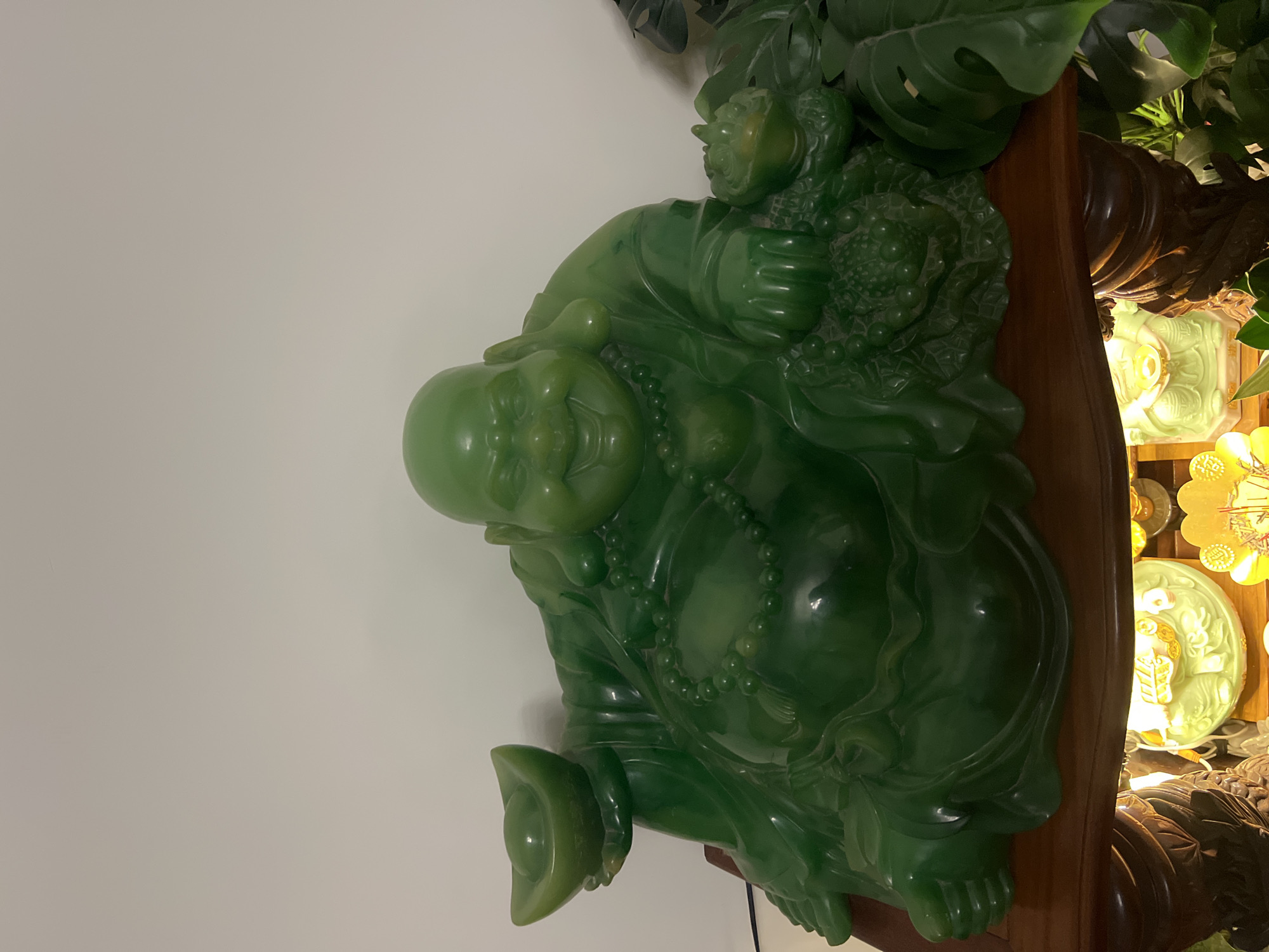 A large, lustrous green statue of Hotei (the "Laughing Buddha" or Budai) is prominently displayed indoors, likely in Bắc Nha Trang, Vietnam. The statue, appearing to be crafted from jade or a similar polished green stone, depicts Hotei with a serene, smiling face, bald head, and a large, round belly. He wears a beaded necklace across his chest.
Hotei holds a cluster of ornate objects, possibly representing fruits or bags of wealth, in one hand, while the other cradles a gold ingot (yuanbao), a traditional symbol of prosperity. The statue is positioned on a dark wooden surface, possibly a shelf or part of an altar.
Behind and to the right of the statue, lush green artificial leaves, resembling tropical plants like Monstera, provide a verdant backdrop. Further to the right, a warm, golden glow emanates from several decorative items, including what appears to be a carved, illuminated plaque or another figure. Above this, a golden coin-like ornament is visible, further emphasizing themes of wealth and fortune. The background on the left is a plain, light-colored wall. The scene suggests a revered display, creating an atmosphere of peace and good fortune. No discernible text is visible in the image.