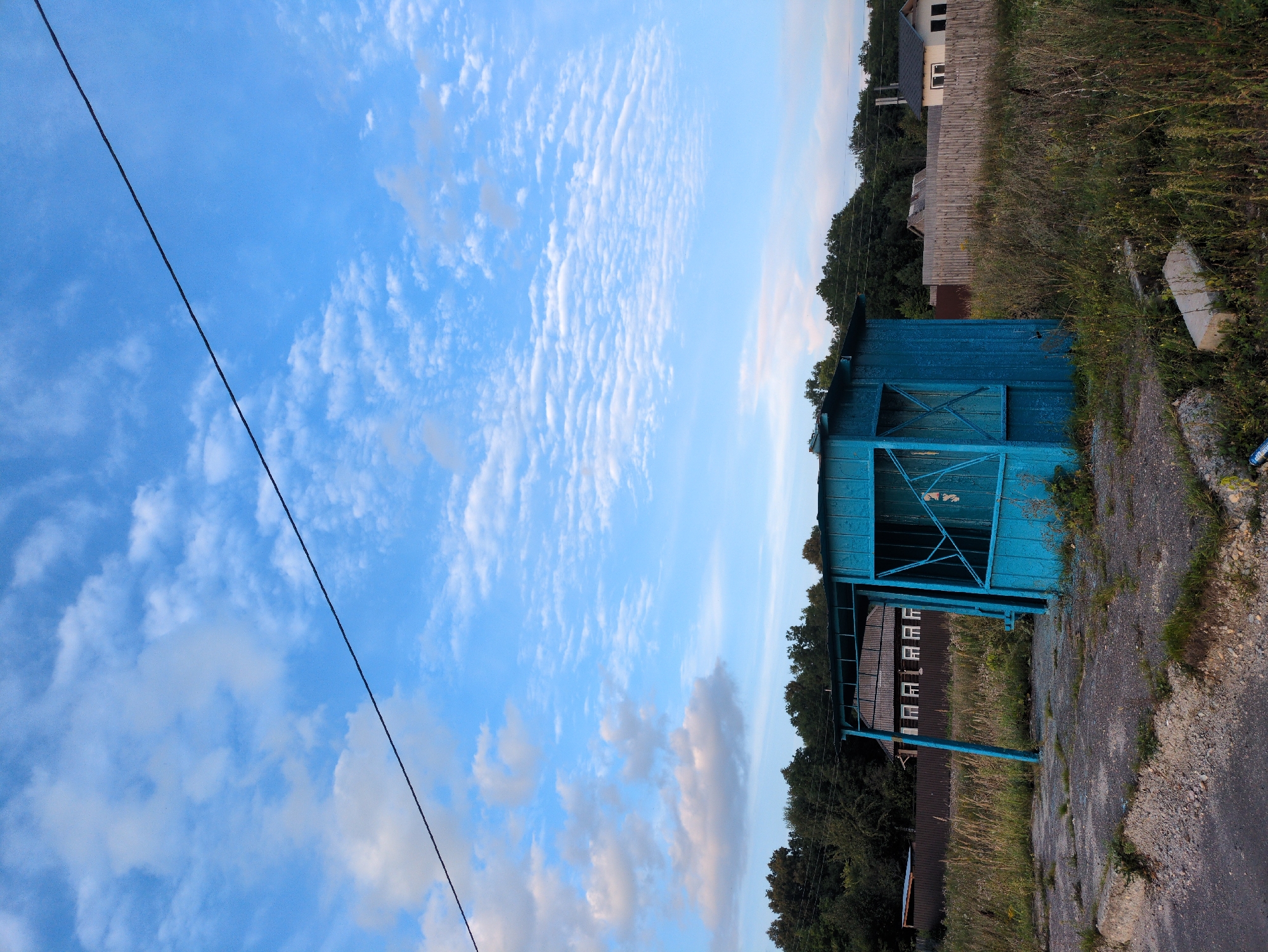 The image depicts an outdoor scene in the village of Posyolok Molochnoy Fermy, Russia. In the foreground, a weathered, bright blue, wooden structure, possibly a bus stop or kiosk, stands in tall, dry grass. The structure has an open front with visible support beams and a simple roof. Paved ground with gravel and rubble surrounds the base of the structure. In the background, a wooden fence separates the area from a residential area with houses and trees. One house is white with a dark roof and windows. The sky above is a vibrant blue with scattered white clouds, some of which are thin and wispy. A power line crosses the upper portion of the frame. The overall impression is of a rural, somewhat neglected setting on a clear day.
