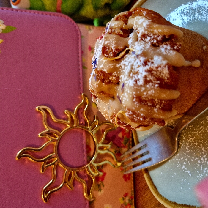 A close-up, high-angle indoor shot presents a single muffin or sweet pastry on a light teal-green plate with a brown rim. The pastry is heavily topped with white icing dripping down its sides, generously dusted with powdered sugar, and adorned with small blue and yellow dried floral pieces. A silver four-tine fork rests on the plate to the left of the pastry, also partially covered in powdered sugar. In the blurred background, a pink handbag or purse is visible, featuring a prominent gold-colored sun emblem with a central circle and radiating wavy rays. The lower portion of the bag displays a floral pattern with red, yellow, and white flowers. To the far right, a green, frog-like plush toy is partially visible. All items rest on a wooden surface. The specific location within the city of Strasbourg, France, is not discernible from the visual content.