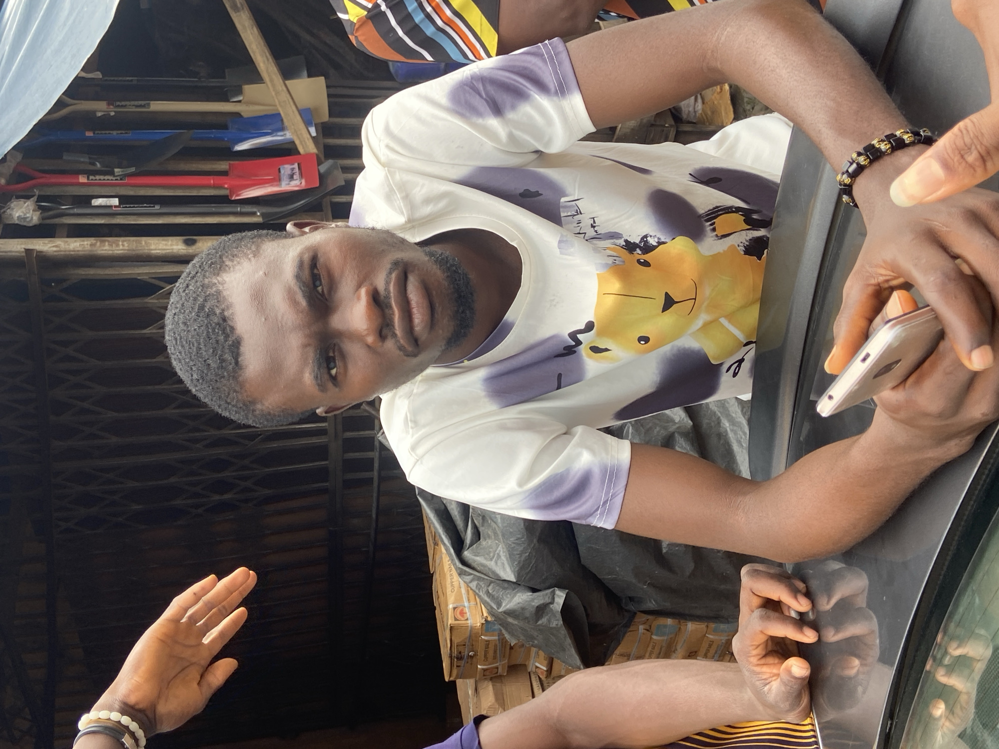 A man sits at the front of a vehicle, his arms resting on the car's surface. He is wearing a white t-shirt with a cartoon bear design. In his hands, he holds a smartphone. To his left, another person's hand is visible, resting on the vehicle. Behind the man, a stack of boxed goods is covered with a black tarp. In the background, several shovels with colored handles hang on a wall. The scene appears to be outdoors in a market or roadside stall, consistent with a location in Ogidi, Nigeria.
