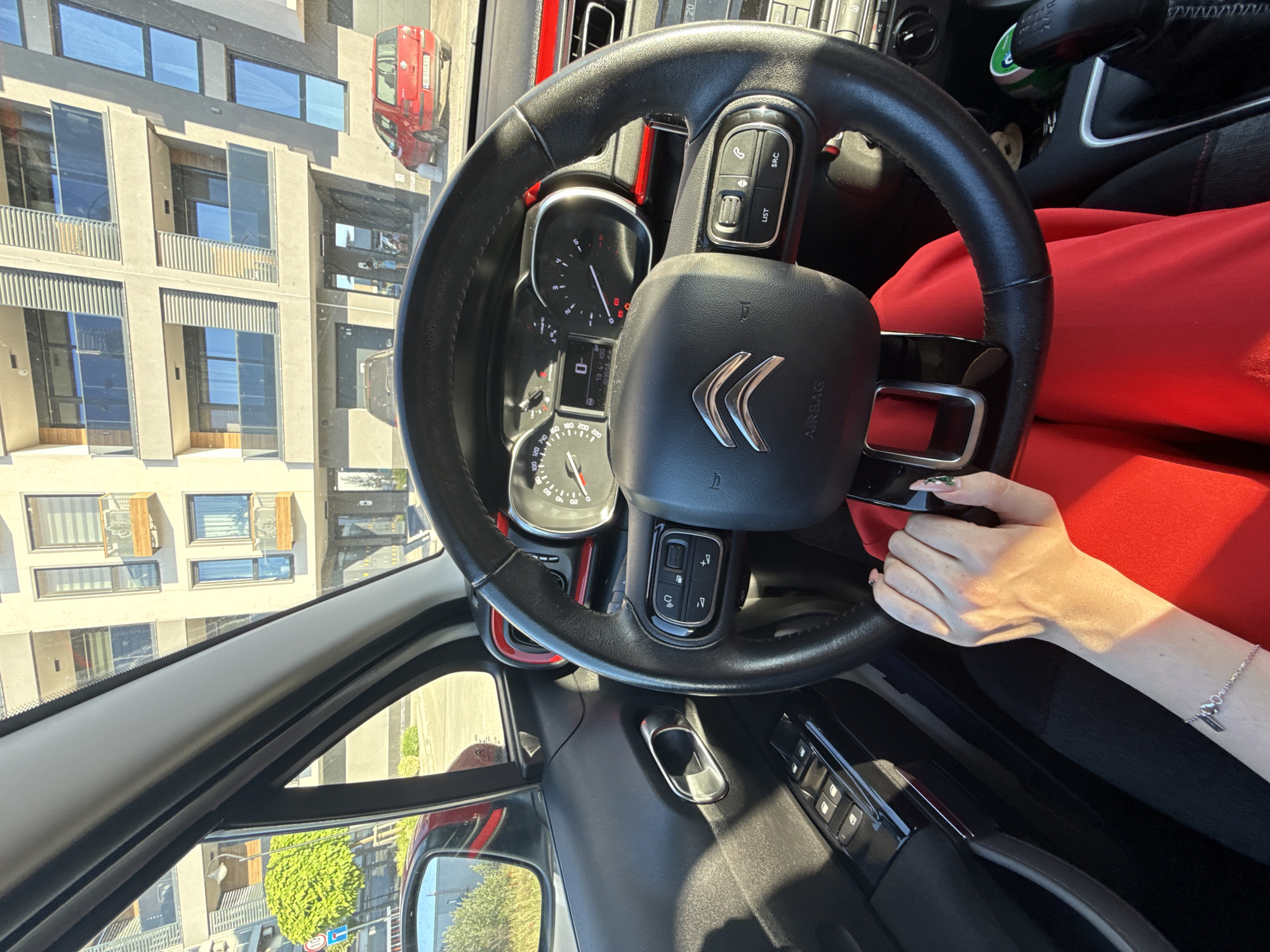 The image shows the interior of a Citroen car, specifically the steering wheel, dashboard, and part of the driver's seat. The steering wheel has the Citroen logo in the center and controls on either side. The dashboard displays the car's instruments, including the speedometer. A person's hand is visible near the steering wheel. The exterior scene visible through the windshield and side windows includes an apartment building, another red car, and some greenery, suggesting a city environment. Based on the vehicle registration numbers partially visible on the red car in the background, the image appears to be in Plovdiv, Bulgaria.