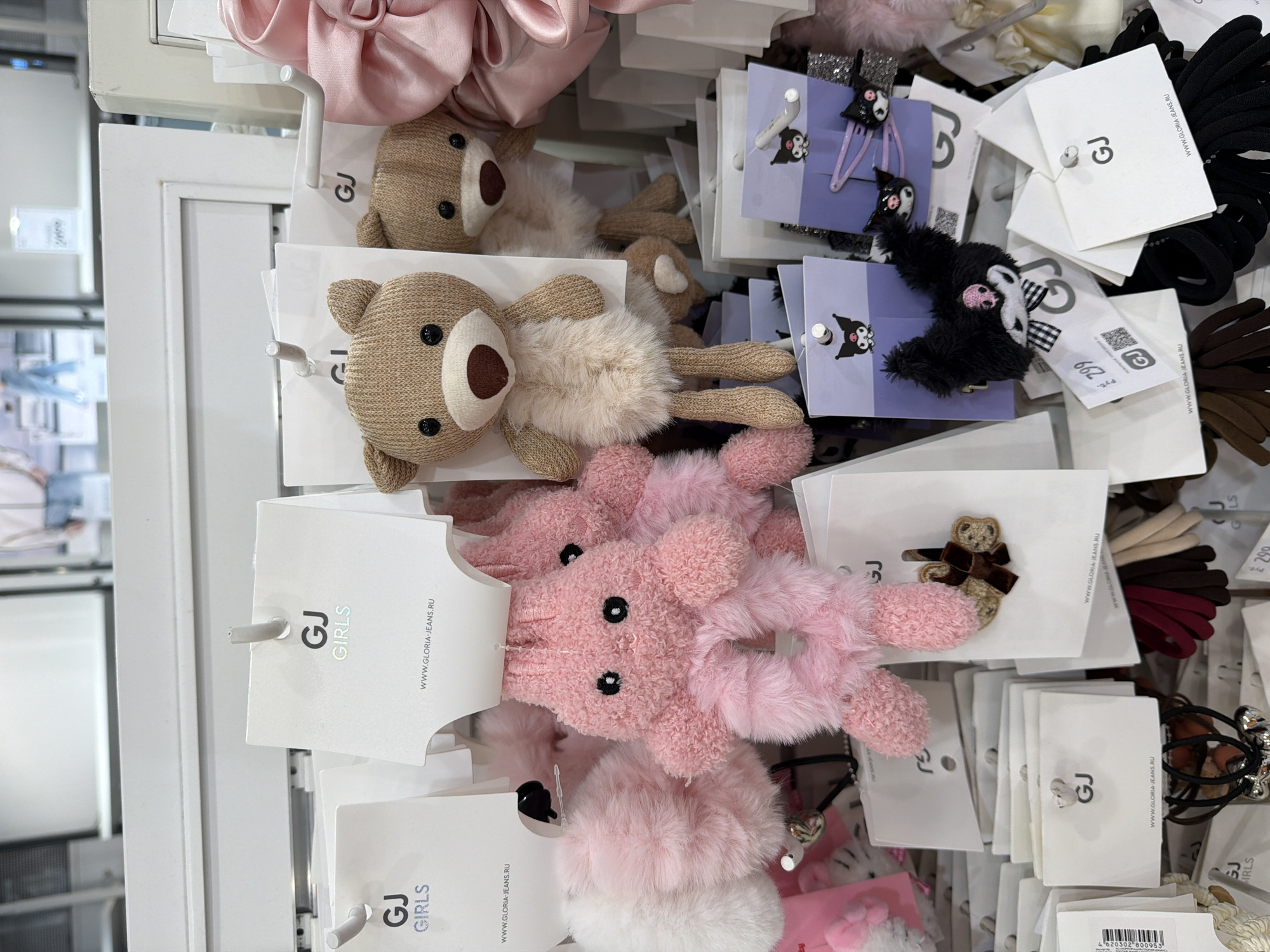 The image depicts a display of hair accessories and plush toys hanging on hooks in what appears to be a retail environment. Various items are visible including hair ties in different colors like black, brown, and burgundy, hair clips with cartoon characters, plush teddy bears, and a pink rabbit-like plush accessory. The items are attached to white tags with the branding "GJ GIRLS" and the website "WWW.GLORIA-JEANS.RU." A tag with the number "299" is also visible, possibly indicating the price. The image suggests a setting in a store like Gloria Jeans, potentially located in Berezniki, Russia, given the website domain.
