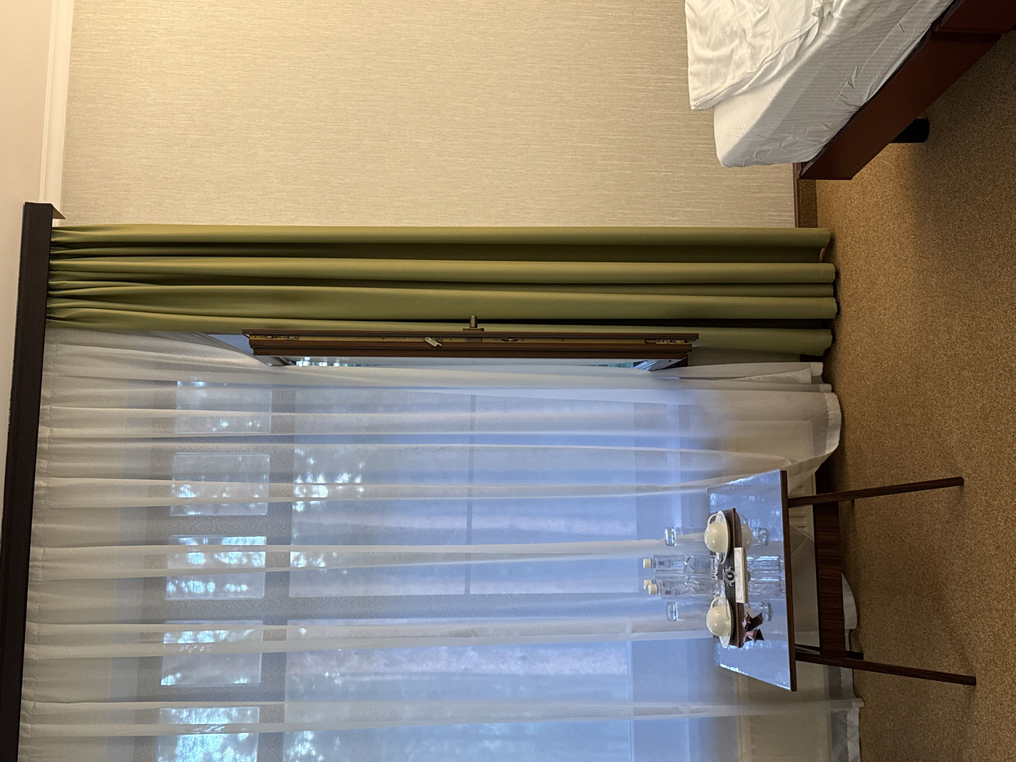 This image captures an indoor scene, likely a hotel room in Журавлево (Кемеровский), Russia. A large window is covered by two layers of curtains: sheer white curtains in the foreground and heavy olive-green curtains to the right. The white curtains reveal a blurry view of trees outside. On a small, dark wooden table in front of the window, there are two cups and saucers, four bottles of water, and two glasses. To the right of the window, a portion of a bed with white linens is visible. The walls are textured in a light beige color. The flooring is a coarse, speckled brown carpet. The scene is lit with artificial lighting, casting a soft glow on the textured wall. There are no people visible in the image.