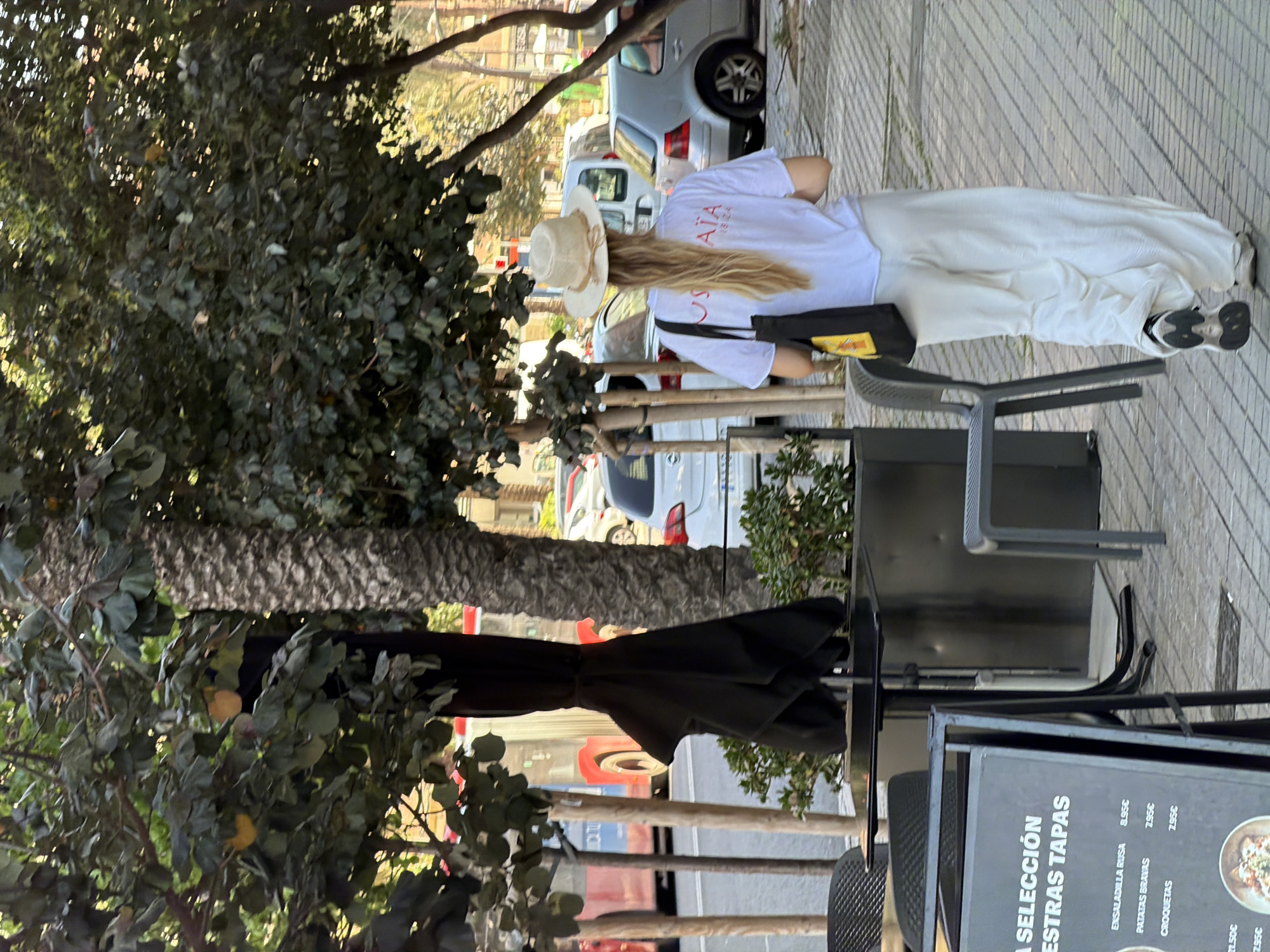 In this full outdoor shot, a woman with blonde hair, wearing a white t-shirt, a straw hat, and flowy white pants, walks away from the camera on a paved sidewalk. She carries a black tote bag with a yellow tag over her left shoulder. The woman walks past an outdoor seating area with black tables and chairs, an umbrella stand, and a menu board. In the background, a red and white bus is visible, along with parked cars and trees. The setting appears to be an urban environment.