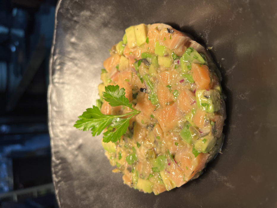 A culinary creation sits on a dark plate, featuring a circular arrangement of diced salmon and avocado. A sprig of fresh parsley adorns the top. The diced ingredients appear to be mixed with a light sauce, adding a glossy sheen. The dish, likely a tartare or ceviche variation, showcases contrasting colors and textures, presented in a simple yet elegant manner.