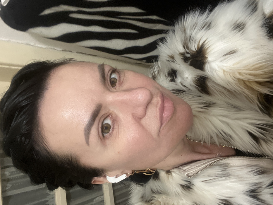 A medium close-up, chest-up portrait depicts an adult female indoors in Smederevska Palanka Municipality, Serbia. The subject, estimated to be middle-aged, has dark, slicked-back hair, light brown eyes, and is looking directly into the camera with a slight smile. She is wearing a cream-colored faux fur jacket featuring irregular dark brown or black spots, with a light-colored zipper visible on the front. A white wireless earbud is present in her left ear, beneath which a gold-toned, abstractly shaped earring hangs. A thin silver-colored necklace with a small pendant is partially visible below the jacket's opening. The background on the left side of the frame shows a light-colored surface with horizontal elements, possibly a window blind or part of a furniture piece. To the right of the subject, a black and white zebra-striped fabric or object is visible.