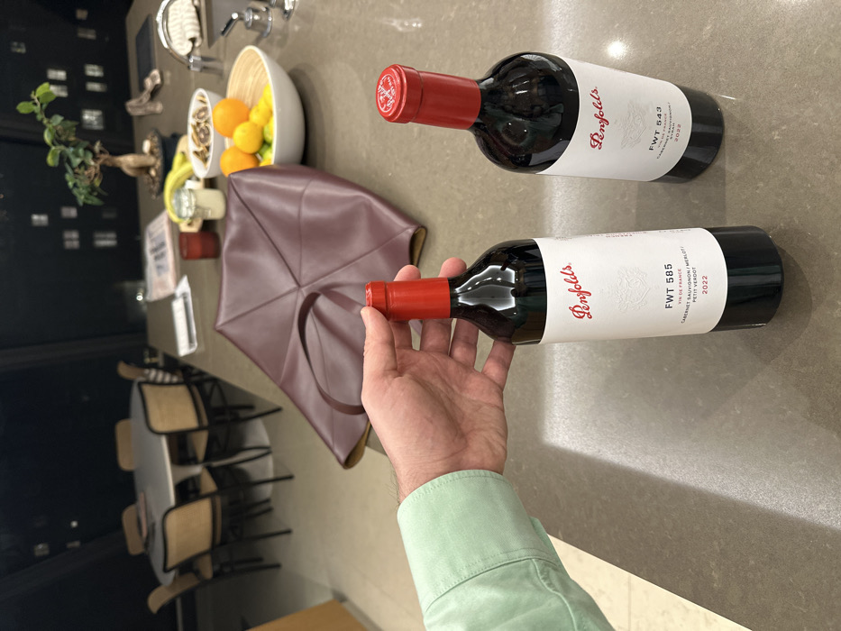 Two bottles of Penfolds wine are placed on a countertop. One label reads "FWT 585, Vin de France, Cabernet Sauvignon/Merlot, Petit Verdot, 2022." The other label reads "FWT 543, 2022". A hand in a light green shirt is holding the "FWT 585" bottle. A burgundy-colored leather bag sits near a bowl of fruit and a decorative tree. A dining table with chairs is visible in the background.