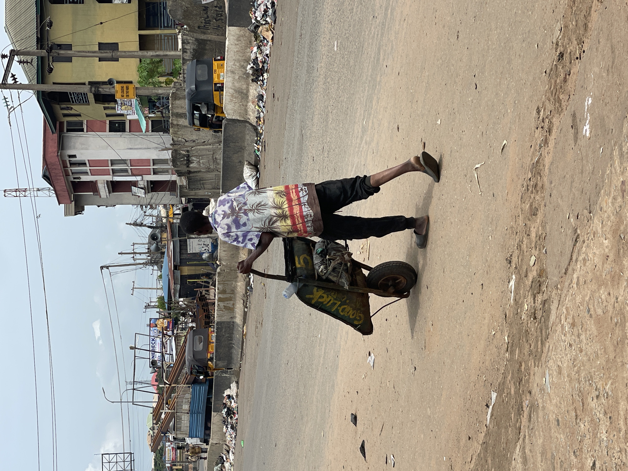 A man pulls a wheelbarrow on a city street in Onitsha, Nigeria. The wheelbarrow is full of objects. The man wears a patterned short-sleeved shirt, dark pants, and sandals. Trash lines the side of the road. Buildings, vehicles, and street signs are visible in the background. Overhead power lines crisscross the scene. The sky is overcast.