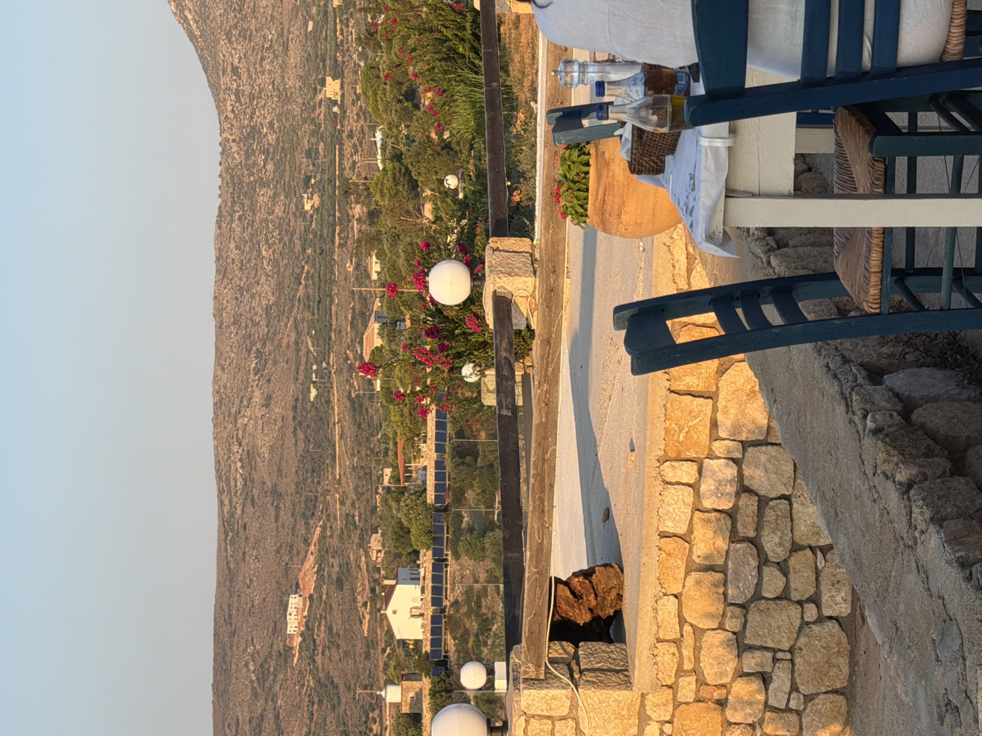 An outdoor restaurant setting is captured in Greece during daylight. The scene overlooks a hilly landscape with scattered white buildings and vegetation. In the foreground, a table is set with chairs, bottles of oil, and a salt and pepper grinder. A large terracotta pot with a plant is also visible. A stone wall separates the restaurant area from the landscape. The sky is clear and blue.