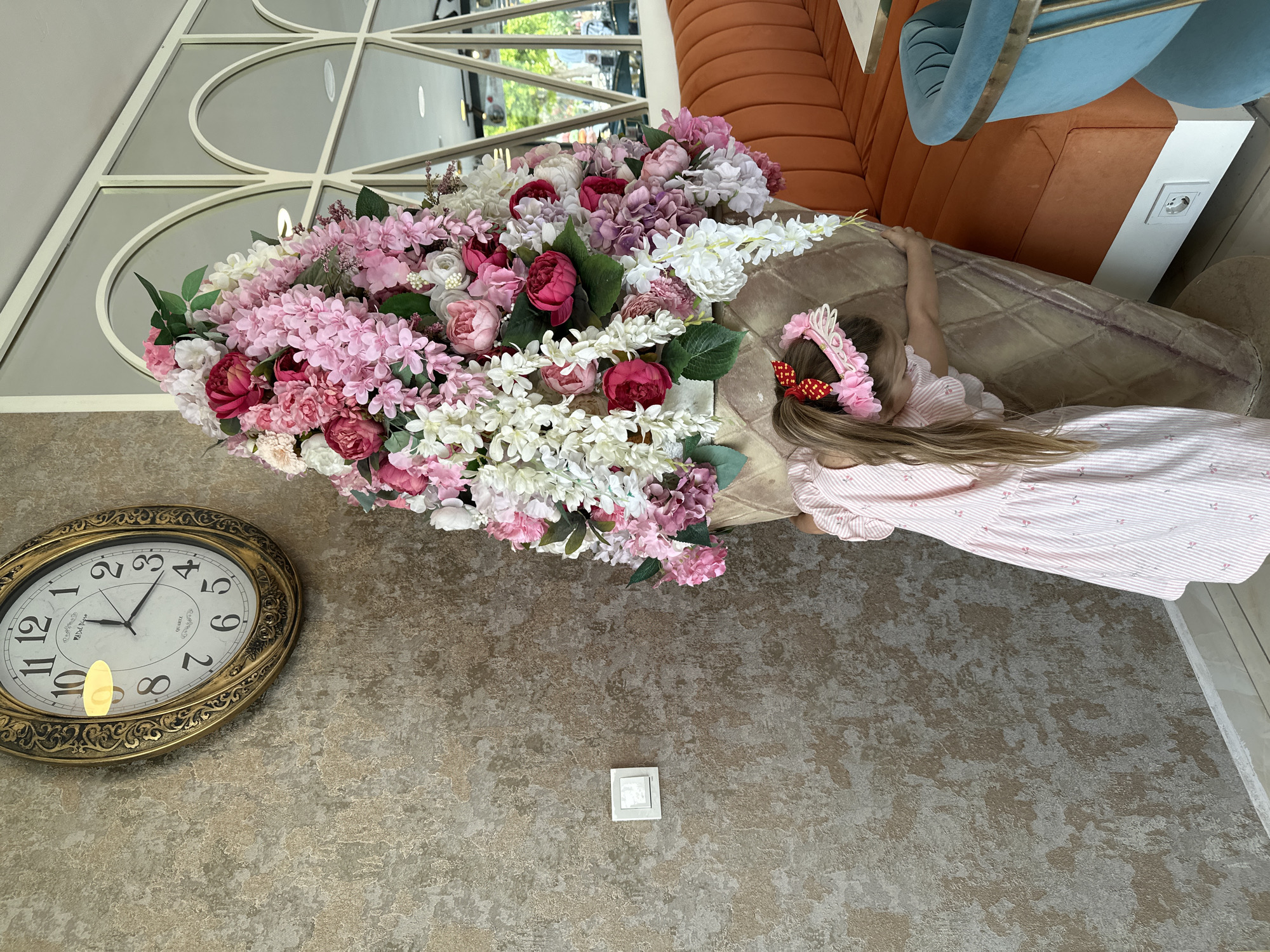 A girl with long blonde hair lies next to an oversized cone filled with flowers. She wears a pink dress with white stripes and a floral headband. The flowers are a mix of pink, white, and burgundy. The cone is tan with a quilted pattern. The scene appears to be indoors, with a wall featuring a decorative clock and light switch. An orange couch and blue chair are in the background, along with a window reflecting outdoor scenery. The location is Fethiye, Turkey.