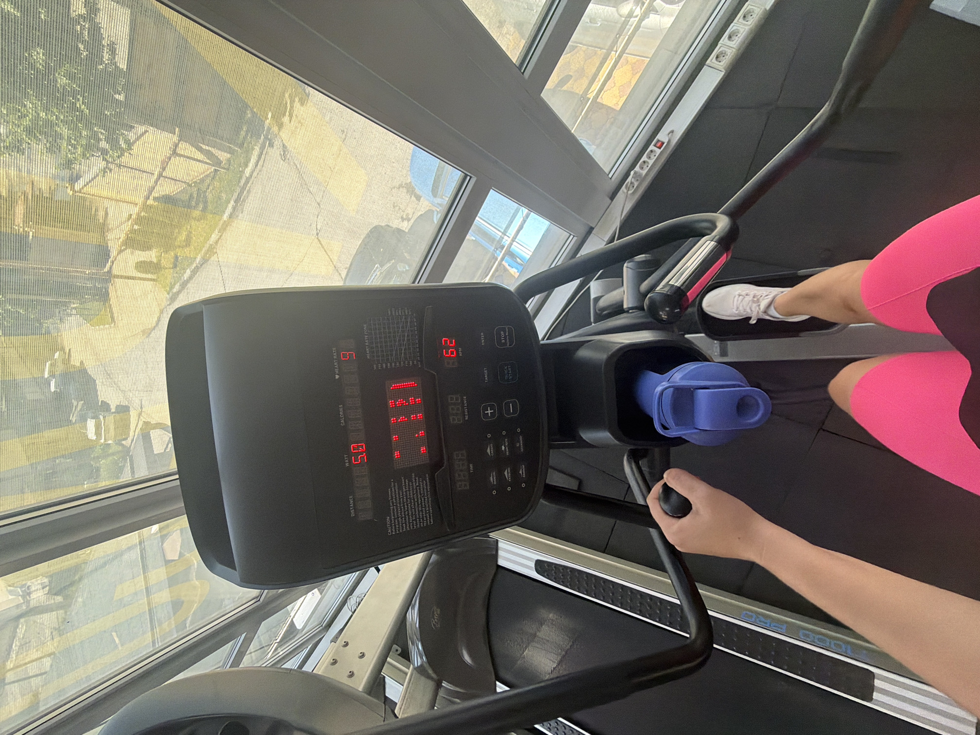 The image captures a person exercising on an elliptical machine within a gym or fitness facility in Sochi, Russia. Bright daylight streams in through large windows that span the length of the room, revealing an outdoor scene with buildings and foliage, partially seen through what appears to be a mesh screen.
A person is visible from the upper thigh downwards, wearing vibrant pink and black athletic shorts and white sneakers. Their left foot is positioned on the elliptical pedal, and their right hand grips one of the moving handles, indicating active use of the equipment. A blue water bottle is securely placed in a holder on the machine's console.
The elliptical's digital display shows various workout metrics. "HEART RATE" reads "9", "RPM" displays "62", and "WATT" shows "5.0". Other numerical sections for "CALORIES", "DISTANCE", and "TIME" show sequences like "14.1.1.8" and "8.8.8.8", though specific values are not fully discernible. Text on the console includes "MODO PRE", "CAUTION", "DISTANCE", "WATT", "CALORIES", "HEART RATE", "RESISTANCE", "+", "-", "TARGET", "HEART RATE ZONE", and a "STOP" button. The model name "1000 PRO" is visible on the side of the elliptical's foot platform. A white power strip with multiple electrical outlets is mounted on the wall beside the window. The scene suggests a focused workout session in a well-lit, modern environment.
