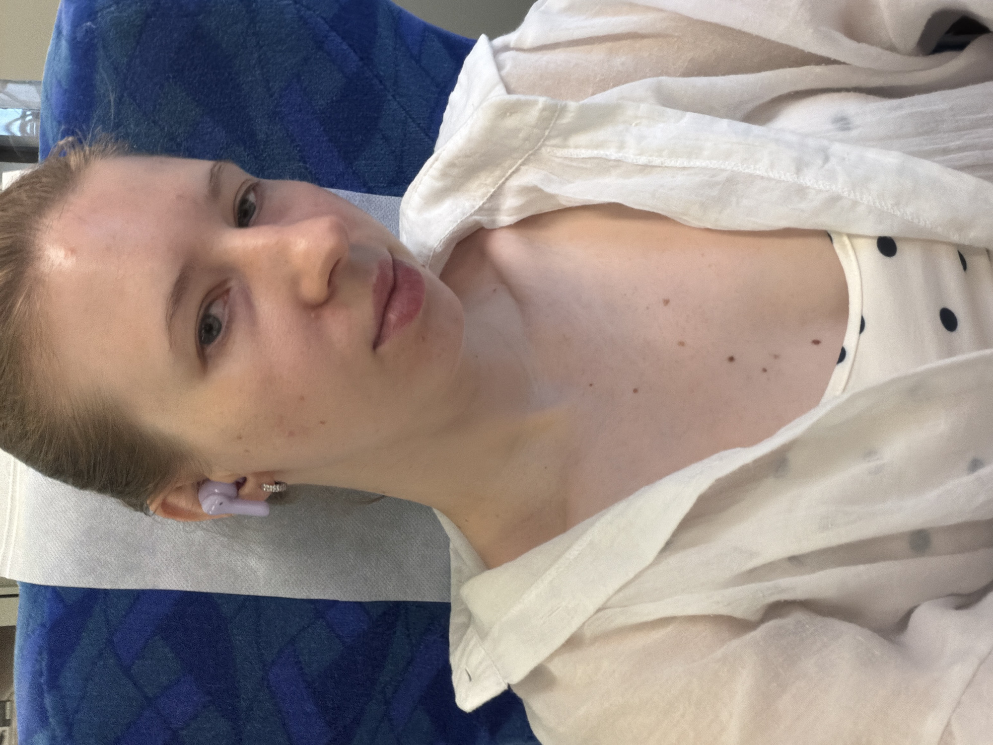 A young woman is seated on a train, likely in Seoul, South Korea, given the context. She is looking directly at the camera, with her head slightly tilted. She has light skin, blonde hair pulled back, and is wearing a white, sheer button-up shirt over a white top with dark polka dots. A light purple earbud is visible in her left ear, and a small hoop earring is in her left earlobe. The background consists of a blue and patterned train seat and a light gray headrest. The lighting is natural and appears to be from an overhead source.