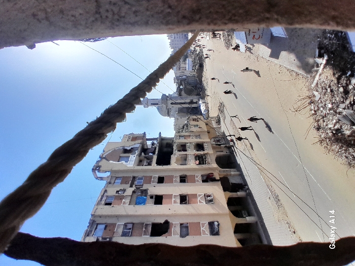 The image presents an elevated view of a heavily damaged urban streetscape in Khan Yunis, Palestinian Territory, under a clear blue sky and bright sunlight. The foreground is partially obscured by two vertical, coarse, cylindrical objects and a horizontal, similar rope-like element, suggesting the photograph was taken from a compromised structure.
On the left, a multi-story building (approximately seven visible floors) exhibits severe damage, with large sections of its facade demolished, exposing internal rooms, blown-out windows, and visible structural damage. Some windows are covered with blue tarps.
The central feature is a paved street, marked with a double solid white line, on which approximately 15-20 individuals are observed walking, casting distinct shadows. The ground immediately bordering the street is unpaved, consisting of sand and debris.
To the right, a substantial amount of rubble and structural fragments are present in the foreground. Further along the street on the right, additional damaged buildings and structures are visible, some partially covered with white tarps.
In the mid-ground, a prominent mosque with a white dome and a tall, slender minaret is visible, also displaying significant structural damage around its base. Beyond the mosque, more damaged buildings are apparent in the distance.