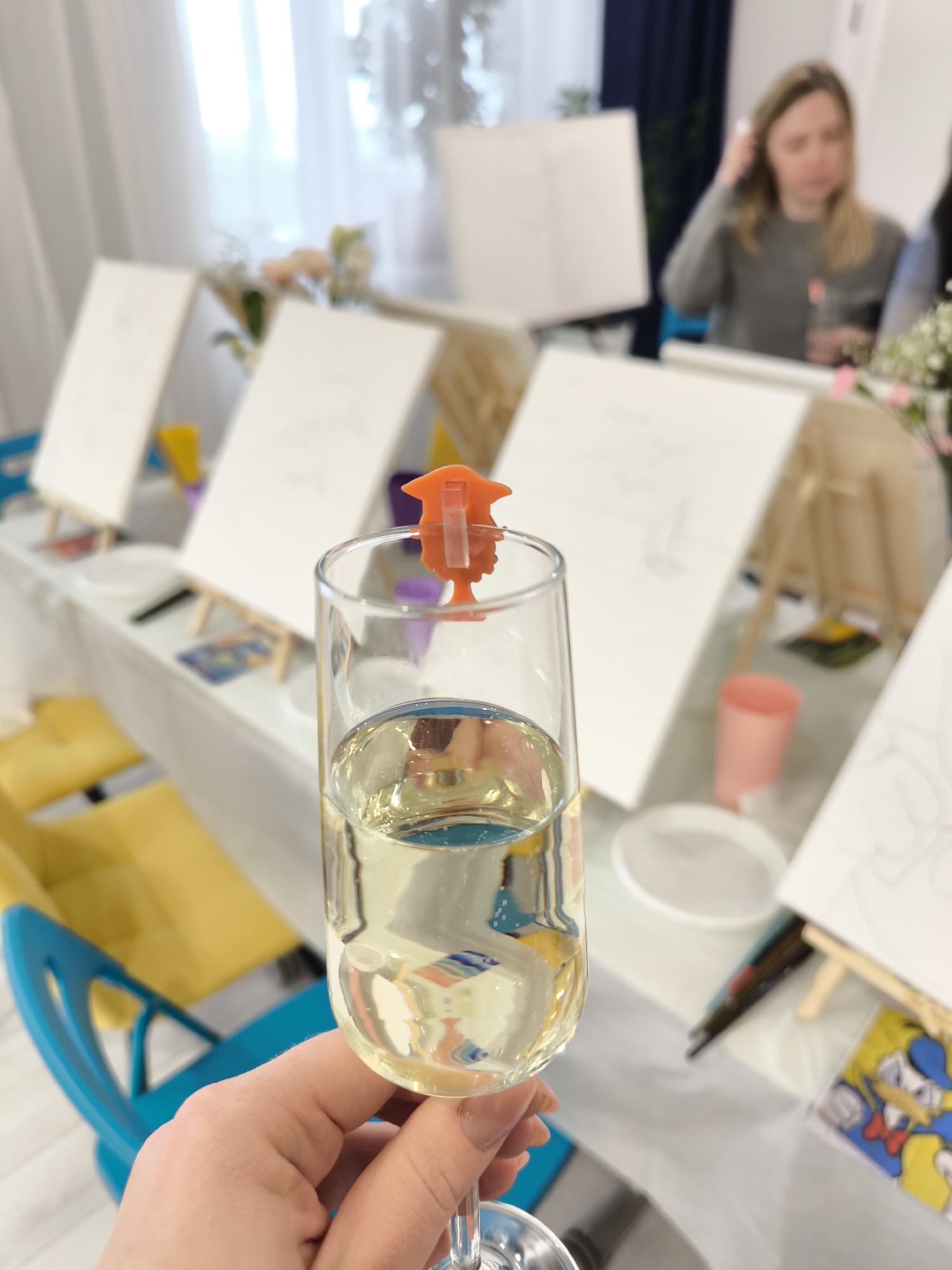 The image shows a close-up view of a hand holding a flute of light-colored sparkling wine or champagne, adorned with an orange decorative graduation cap on the rim. The background reveals a brightly lit indoor setting suggestive of an art class or party, with multiple easels bearing white canvases, likely at a paint-and-sip event. There are flower arrangements on the tables, along with art supplies and various cups. One woman can be seen in the background, possibly engaged in conversation or setting up. The location is reported to be in Fryazino, Russia.