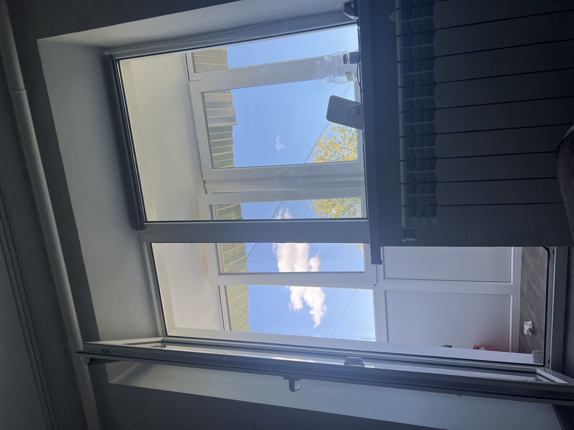 The image depicts a view from inside a room, looking out through a large, opened window and a partially open adjacent door. The setting appears to be an apartment or residential building in the city of Sheptytskyi, Ukraine.
The window system is modern, with white PVC frames. The main window area shows a bright blue sky with scattered white clouds. Through the windows, trees with yellowing foliage are visible, suggesting it is likely autumn. Outside the windows, a light green awning or roof structure is visible overhead.
In the foreground, partially obscuring the lower part of the window, is a white radiator with vertical fins. On the windowsill, there are several objects: a white mobile phone in a stand, a clear glass jar, and a small bottle with a red cap.
The opened door reveals a small balcony or an adjacent enclosed space with white walls and a wooden floor. A crumpled piece of white paper is on the floor near the doorway.
The overall lighting suggests a daytime scene, with sunlight streaming in. The room interior is dimly lit, creating a contrast with the bright outdoor view. The image is taken from a slightly low angle, looking upwards towards the window. There are no people visible in the image.