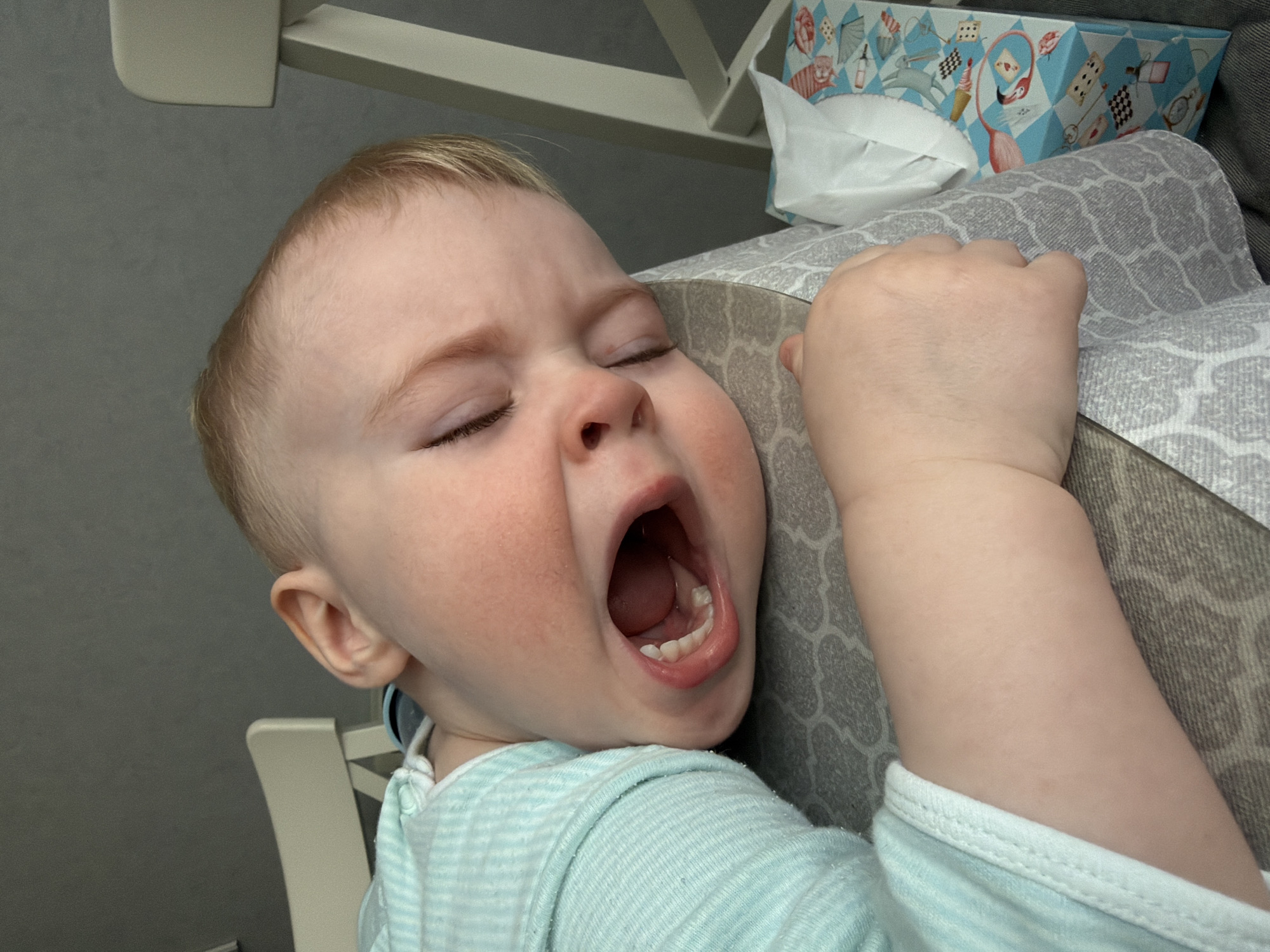 A close-up image shows a young child, approximately toddler-aged, with fair skin and light hair, in Votkinsk, Russia. The child's head is tilted to the right, resting on a gray surface with a light geometric pattern. The child's eyes are closed, and the mouth is open wide, revealing developing teeth. The cheeks appear flushed. The child is wearing a light blue and white striped long-sleeved garment. To the child's right, a rectangular tissue box with a light blue background featuring multi-colored illustrations and a white tissue protruding is visible. Partial views of two light-colored chairs and a plain dark gray wall are in the background.