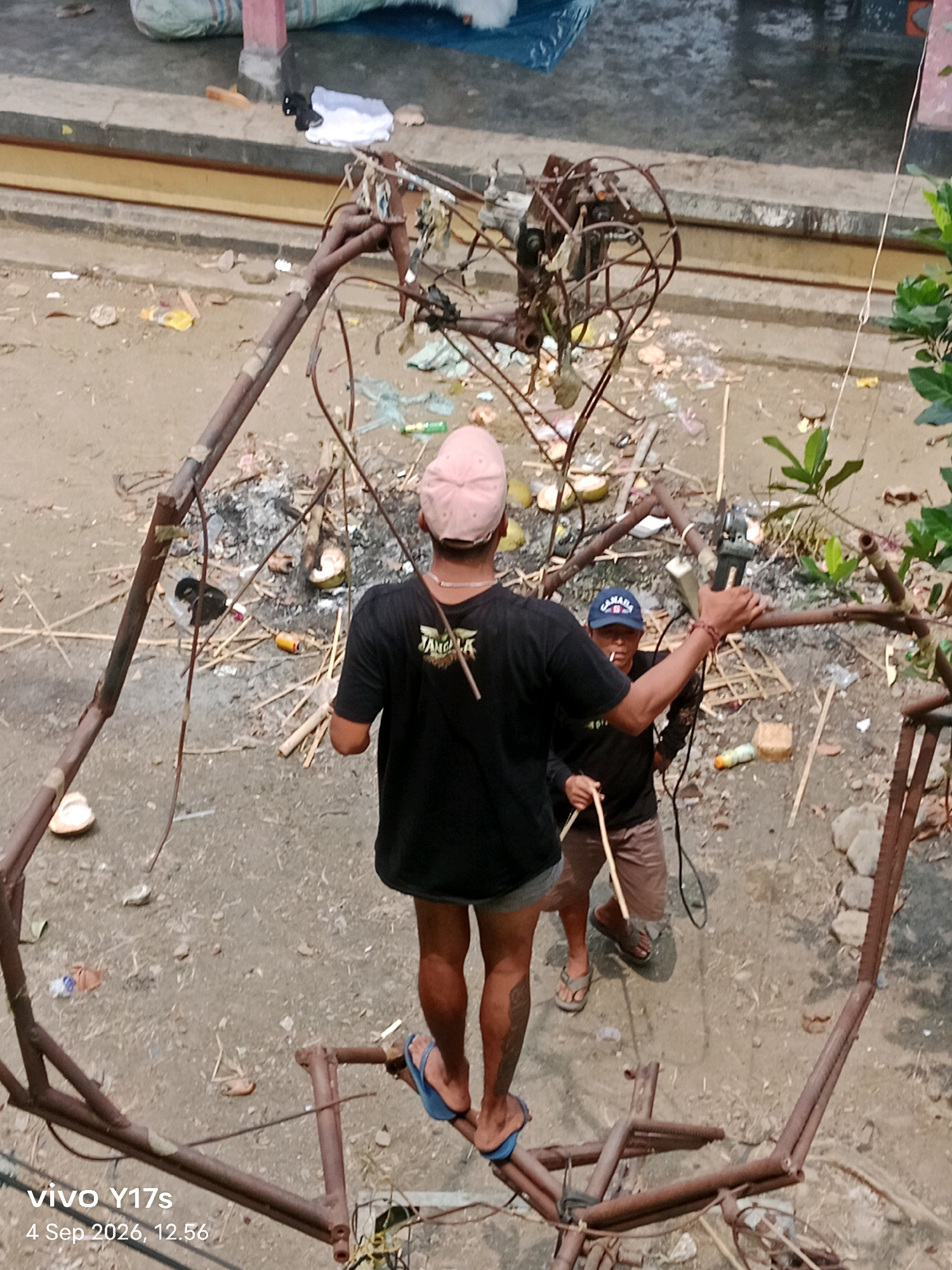 In Rama Dewa, Indonesia, two men are engaged in work on a metal structure. The man in the foreground, wearing a pink cap and a black t-shirt, stands on a metal beam, his right leg extended. He appears to be holding a tool or object in his right hand. In the background, another man, wearing a blue baseball cap and a black shirt, holds a tool that resembles a grinder, aiming it at the metal structure. Both men are dressed casually. The environment is an outdoor area with a dirt ground covered in debris, including bamboo sticks, plastic waste, and discarded items like coconut shells and cans. The metal structure itself is constructed from rebar and pipes, appearing unfinished or partially dismantled. In the upper background, a tiled ledge with some fabric items is visible, suggesting a nearby building or structure. The overall scene depicts an industrial or construction-related activity in an unkempt outdoor setting.