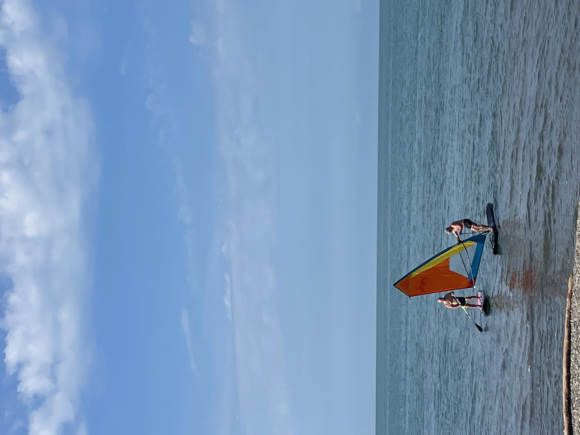 Two men are windsurfing in the calm, teal waters of the Black Sea, near the city of Sochi, Russia. The sky above is a bright, cloud-dappled blue. The man on the left is standing on a paddleboard, holding a paddle, and is positioned to the left of a colorful windsurfing sail. The man on the right is standing on a windsurfing board, actively engaged in windsurfing. The sail is triangular and features sections of orange, yellow, and blue. The water is relatively still, with gentle ripples. In the foreground, a shoreline of pebbles and a piece of driftwood are visible.