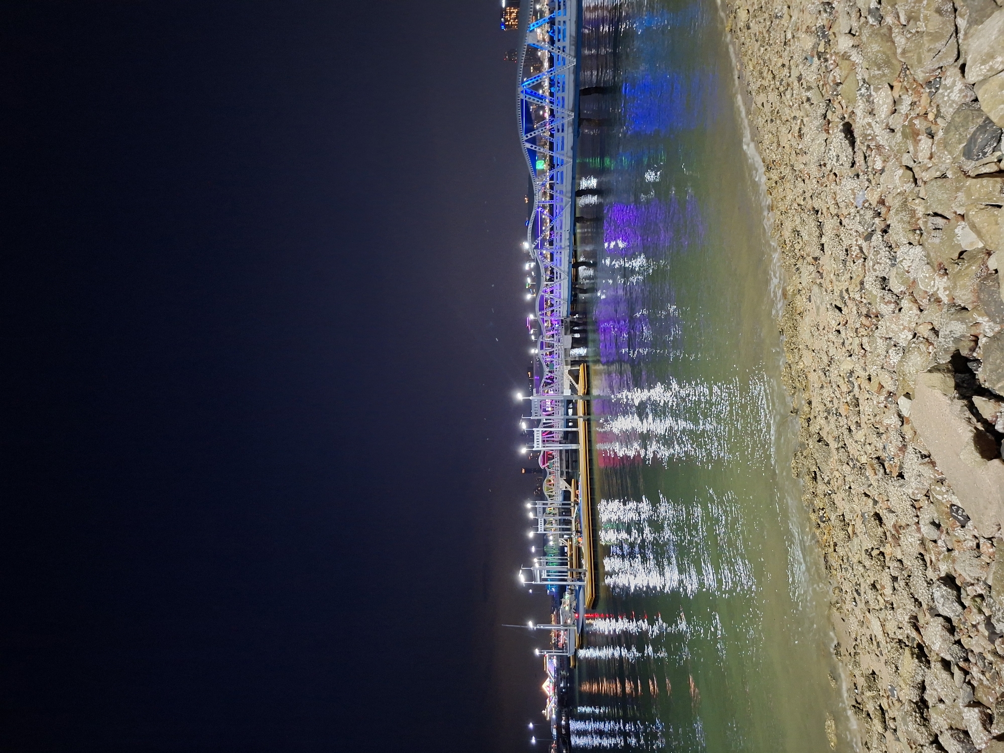 This nighttime image captures a coastal scene in Pattaya City, Thailand. A bridge adorned with colorful lights stretches along the coast, its vibrant reflections shimmering on the water's surface. The shoreline consists of a mix of sand and rocks, while the background is dominated by a dark, starless sky. The water is calm with clear reflections of the lights.