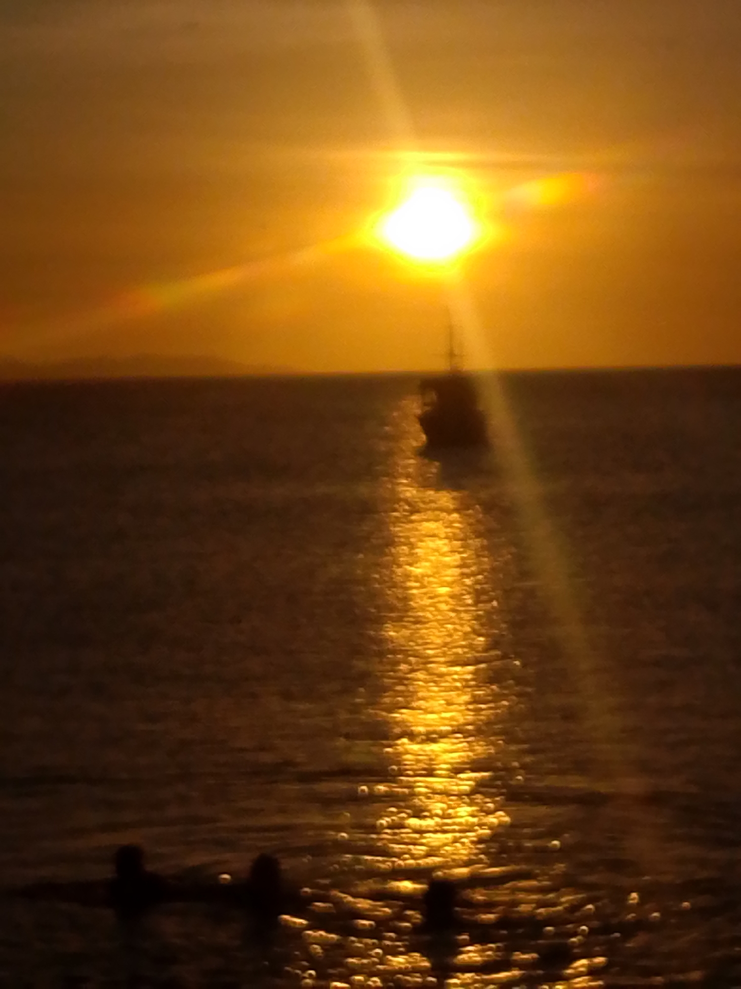 This image captures a sunset scene over a body of water in Juan Griego, Venezuela. The upper portion of the frame is dominated by a brilliant, white-hot sun positioned centrally, casting intense yellow and orange light across the sky and generating prominent lens flares. A dark, indistinct landmass is visible along the distant horizon line. The lower two-thirds of the image consist of a dark body of water, across which a shimmering, vertical path of golden light extends directly from the sun towards the foreground. In the mid-distance, a dark, silhouetted boat, appearing to be a sailboat with a visible mast, is present on the water. In the lower-left foreground, three dark, indistinct human figures are partially submerged in the water, visible as silhouettes. All foreground and mid-ground subjects are rendered in deep shadow due to the strong backlighting from the sun.