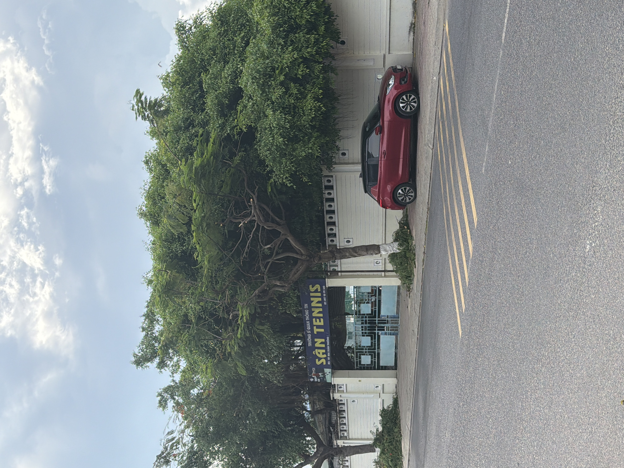 The image depicts the entrance to a tennis court facility in Bac Nha Trang Ward, Vietnam. A red hatchback car is parked on the sidewalk to the right of the entrance. The entrance itself features a blue banner with yellow text that reads "SÂN TENNIS" and "TRƯỜNG SĨ QUÂN THÔNG TIN". Behind the entrance gates, some blue structures are visible. A white fence with decorative circular patterns lines the property. The area is bordered by a paved road with yellow pedestrian crossing markings. Lush green trees, some with visible branches, overhang the entrance and the fence. The sky above is partly cloudy with a light blue hue.