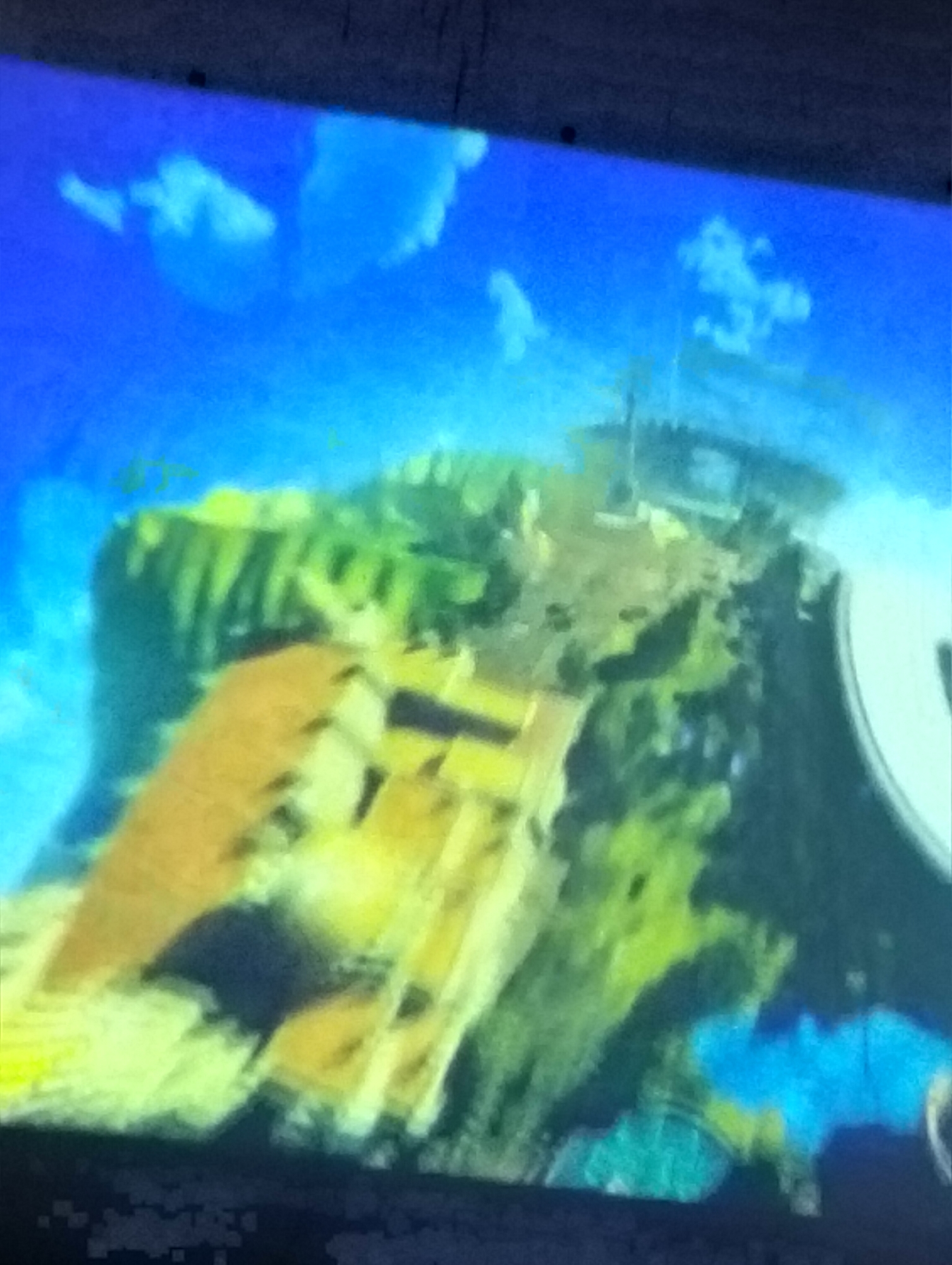 The image shows a blurry, pixelated representation of a vibrant, cartoon-like landscape. The scene depicts a lush, green, and brown cliffside with a bright blue sky dotted with white clouds. There appears to be some sort of structure or pathway on the cliff, possibly a ramp or staircase, rendered in shades of yellow and brown. The overall impression is of an animated or video game environment. There are no discernible people, objects, or specific actions taking place in the image. The location in the city of Payagaboh, Indonesia, is not identifiable from the provided visual information.