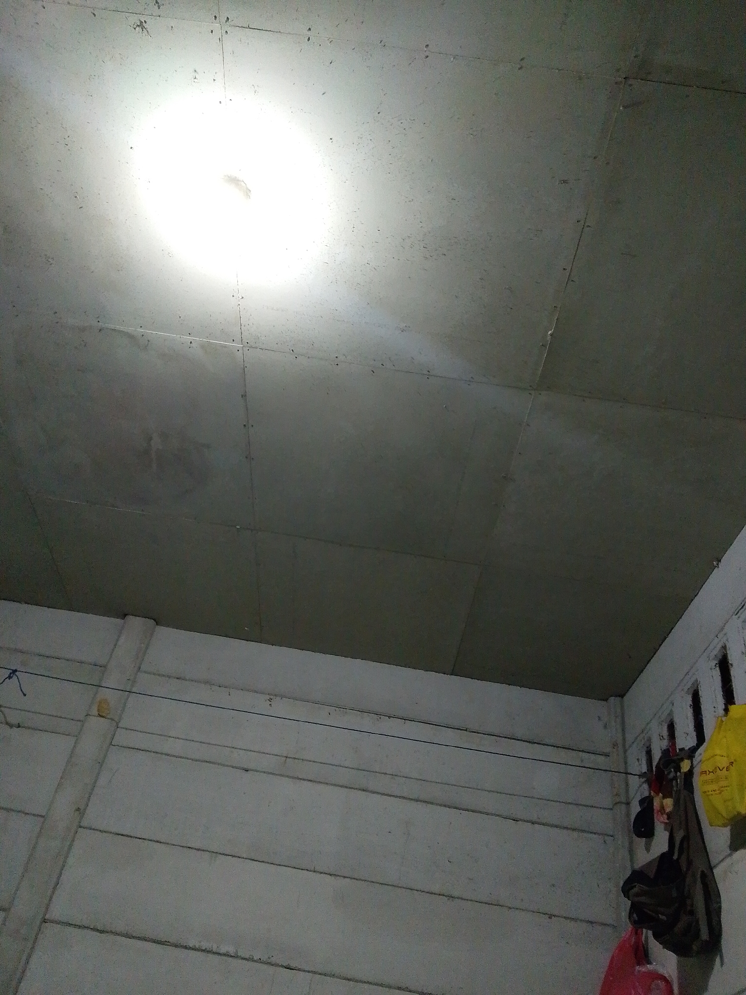 The image depicts an interior scene in Bukit Ketok, Indonesia. The ceiling is constructed of square concrete panels. A bright light source, likely a bulb, is mounted centrally on the ceiling. The walls are built of horizontal concrete panels, possibly pre-fabricated sections. Clothes are hanging from a line attached to a section of wall with vertical slits on the right side of the image. There are several bags of various colors hanging on the wall near the clothes.
