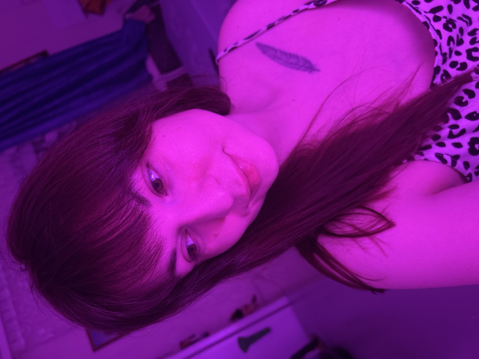 A woman with long brown hair and bangs is lying down, viewed from above and angled. She is wearing a top with a leopard print pattern. A tattoo of a feather is visible on her chest/shoulder area. The lighting is predominantly purple. The background includes what appears to be a blue curtain and objects on a surface. There is no indication of a location in the city of Фоки, Russia.
