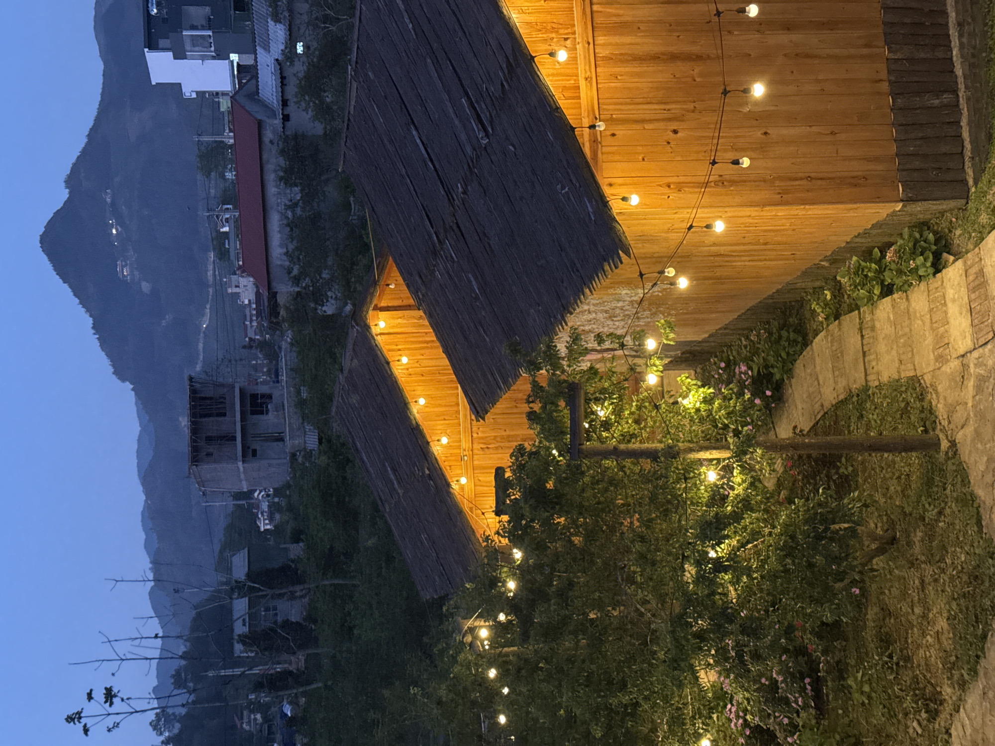The photograph depicts an outdoor scene in Bắc Hà, Vietnam, at dusk. A cluster of thatched-roof structures, possibly part of a restaurant or homestay, is illuminated by strings of small, warm-toned lights. Wooden walls and walkways are visible. Dense foliage and greenery fill the foreground and midground, partially obscuring the structures. In the distance, a mountain range and residential buildings are silhouetted against the evening sky. The image captures the atmosphere of a rural or semi-rural location with a mix of natural and built elements.