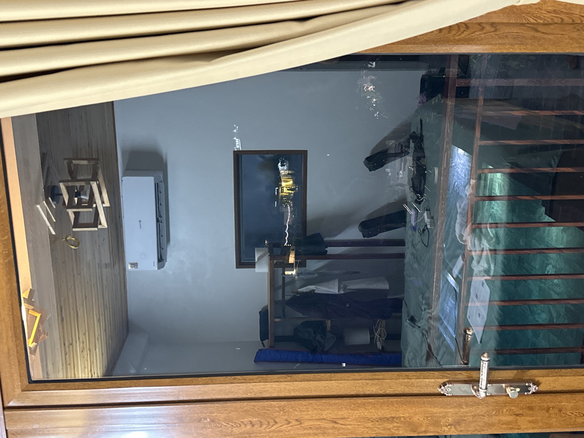 The image shows a bedroom viewed through a glass door from the exterior. The room appears to be located in Xóm Khán, Vietnam.
Inside the room, a bed with a green duvet is visible. There is a shelf unit with clothes hanging on it, including what appears to be a dark coat and a white shirt. On the shelf, there are several items, including a blue cylindrical object, a black bag, and a white roll, possibly of toilet paper. Above the shelf, there is an air conditioning unit mounted on the wall.
A window is present on the wall, through which a reflection of outdoor lights and buildings can be seen. The ceiling is made of wooden planks. A wooden chandelier with a geometric design hangs from the ceiling.
The glass door has a wooden frame and a metal door handle. A cream-colored curtain is visible to the right of the door, obscuring part of the interior.
The overall impression is of a residential or hotel room, likely in a town or city given the reflected lights in the window.