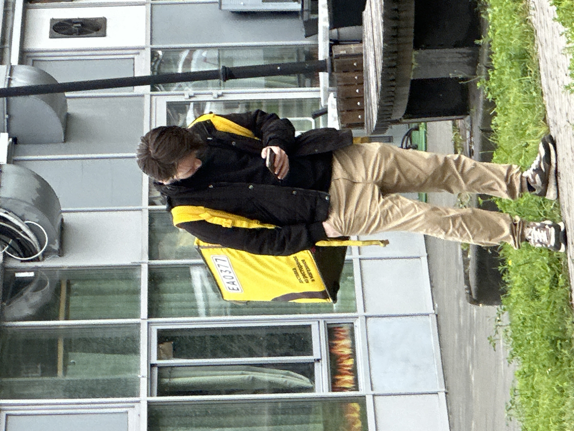 A male courier wearing a black jacket with yellow accents and khaki pants stands on a paved surface next to a grassy area. He has a yellow delivery backpack on his back, which has "EA0377" printed in black on a yellow rectangle, and text in Russian below that reads "доставка из ресторанов и магазинов" (delivery from restaurants and shops). The courier is looking down at his mobile phone, which he holds in his right hand. He is wearing dark sneakers with white soles. Behind him is a modern building with large windows, some of which have a green tint. Visible in the windows is a reflection of flames. In the background, part of a dark lamppost is visible, as well as a section of a large, circular wooden structure that appears to be a table or seating area. The overall environment suggests an urban setting, possibly a commercial or residential area in Podolsk, Russia.