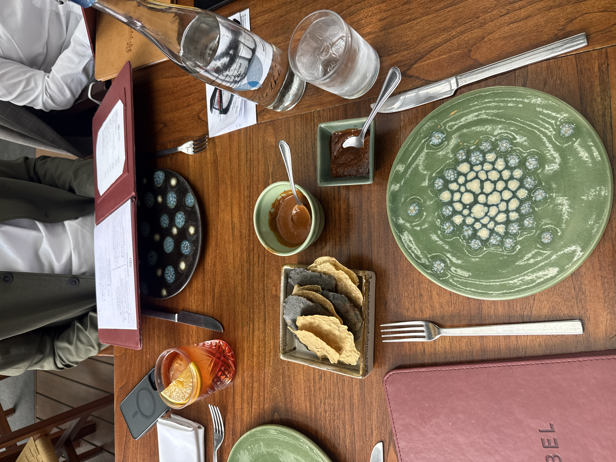 A high-angle shot shows a table at Contramar, a seafood restaurant in Mexico City, Mexico.
Subjects: Two individuals are partially visible. One, on the left, wears a white shirt and a green jacket. Another individual has only an arm visible, clad in a white shirt and gray suit jacket.
Objects: The table is covered with a dark wood surface. Objects include two menus (one red and one burgundy), two decorative plates (one green and one black with blue patterns), two small bowls of sauces, a glass of water, a bottle of water, a glass of cocktail with orange, a phone, a stack of chips in a rectangular holder, several forks and knives, napkins, and placeholders.
Actions/Interactions: The focus is on the arrangement of objects, seemingly before or during a meal. The positioning of the menus suggests that food selection might be in progress.
Environment: The setting is a restaurant. Contramar is known for its seafood and stylish atmosphere.