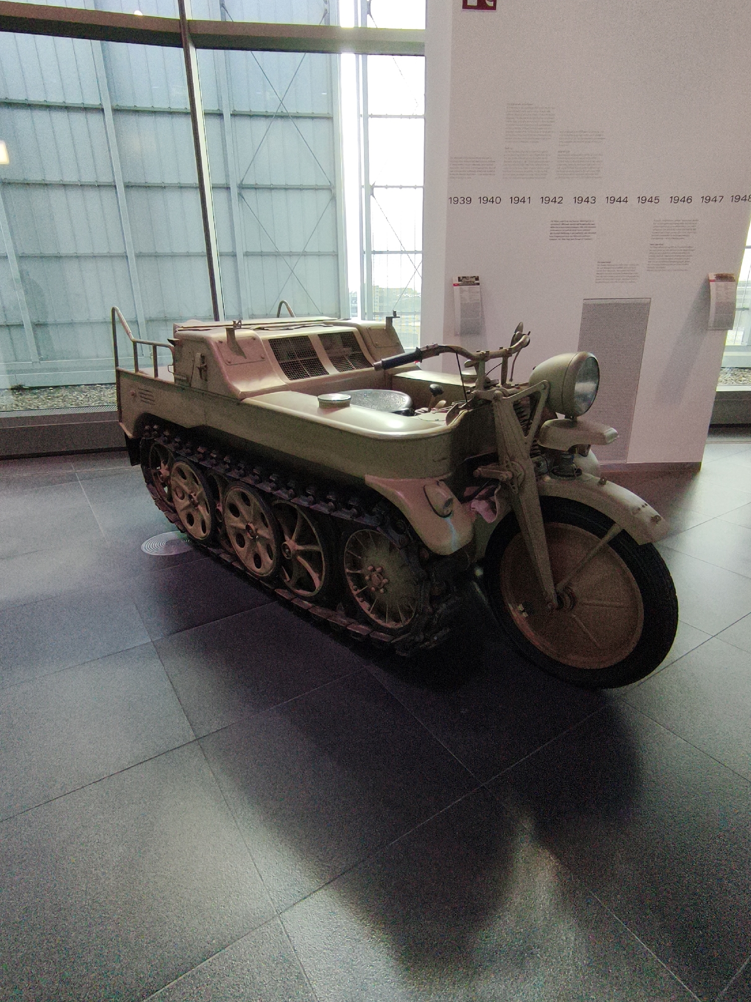 The image displays a Sd.Kfz. 2 Kettenkrad, a half-track motorcycle used by Germany during World War II, as an exhibit in a museum. The Kettenkrad is light tan in color and features a front motorcycle wheel and rear tracks with several smaller road wheels. It is positioned on a dark, reflective floor. In the background, a wall with a timeline marked with years from 1939 to 1948 is visible, suggesting the museum's focus on a specific historical period. The environment appears to be an indoor exhibition space with large windows and metal framing in the upper left corner. The lighting is typical of a museum setting, with spotlights and ambient light creating reflections on the floor and the exhibit. The scene is static and shows no people. The museum is the Audi Forum Ingolstadt, located in Ingolstadt, Germany.