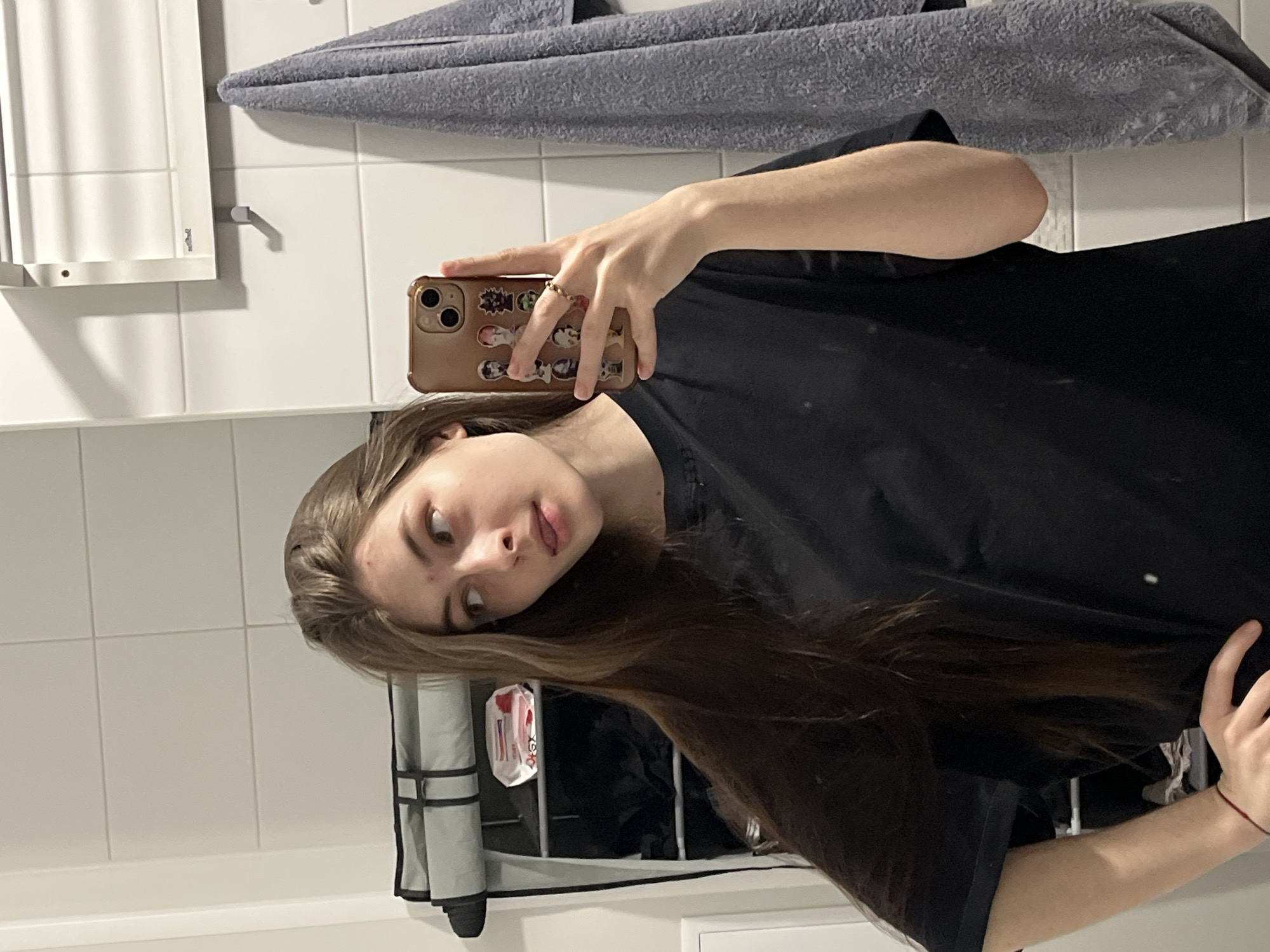 A young woman is taking a selfie in a bathroom. She is holding a brown smartphone with stickers on the back. She has light skin, brown hair, and is wearing a black t-shirt. The bathroom has white tiled walls, a gray towel hanging on the wall, and a toiletries rack. The image provides no information to determine a location in Saint Petersburg, Russia.