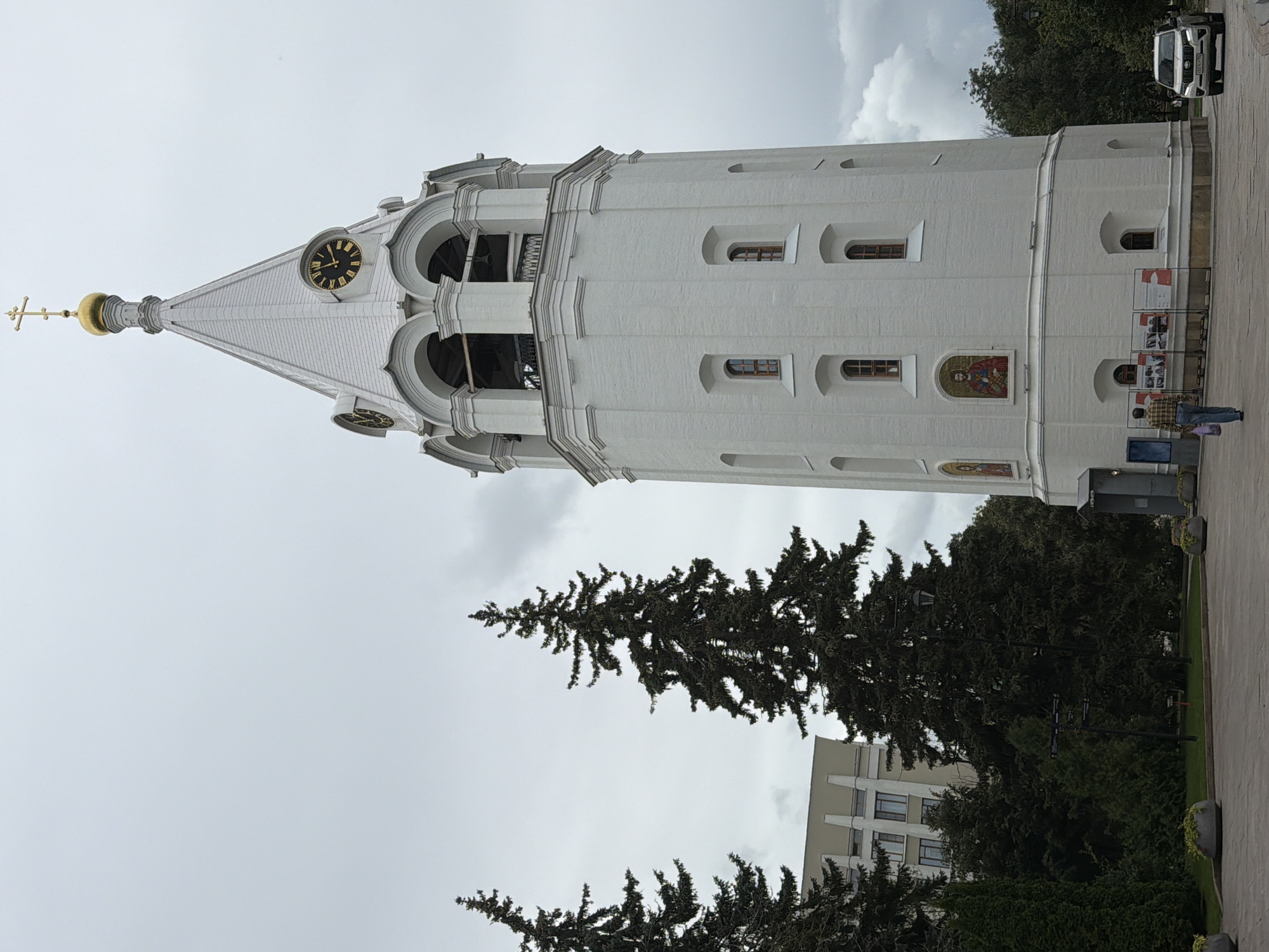 The image shows the Dmitrovskaya Tower in Nizhny Novgorod, Russia, under an overcast sky. The white, brick tower, a prominent landmark, has a golden dome topped with a cross. A clock face is visible on one of its upper sections. Two tall fir trees stand to the left of the tower, with a modern building in the background. In the foreground, a paved area is visible with some people walking and a white SUV parked on the right. There are also some display boards in front of the tower.