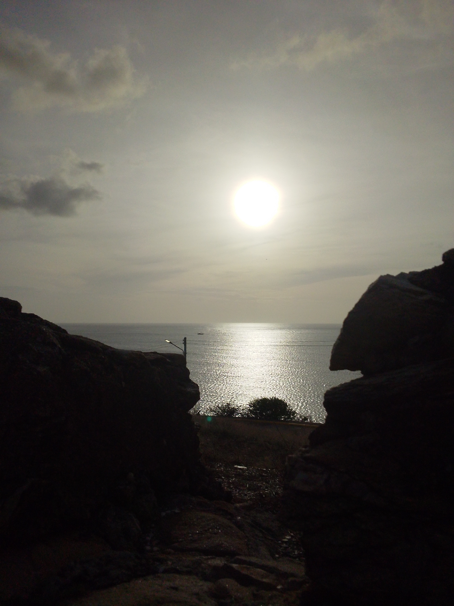 The image captures a view of the ocean from a rocky outcrop in Juan Griego, Venezuela. The sun is prominently visible in the sky, casting a bright glare on the water's surface, which shimmers with reflected light. In the distance, a small boat can be seen on the ocean. The foreground is dominated by dark, silhouetted rock formations that frame the scene. A single lamppost is also visible on the edge of the cliff, suggesting the location is near a pathway or overlook. The sky is overcast with scattered clouds, but the sun's intensity is still strong. A small clump of trees is present on the land between the rocks and the water.
