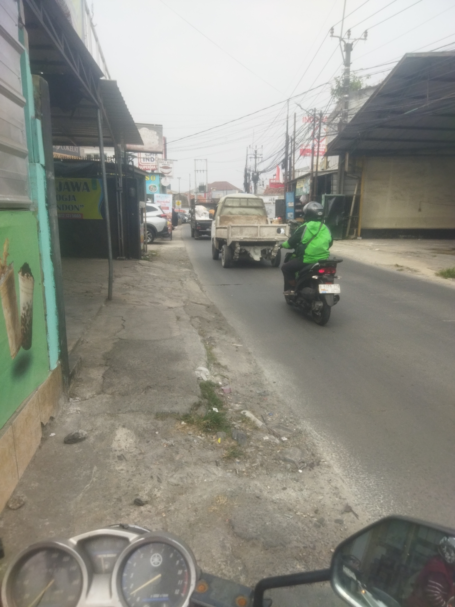 The image depicts a street scene in Depok, Indonesia, under an overcast sky. The perspective is from a moving vehicle, likely a motorcycle, with the dashboard visible in the foreground.
On the left side of the road, a building with a green facade and a corrugated metal awning is visible. A sign on the building reads "JAWA", "OGJA", and "NDON". There are also depictions of drinks on the green wall.
The road itself is paved and appears to be a two-lane street with traffic. A person wearing a green jacket and a helmet is riding a motorcycle in the right lane, moving away from the viewer. Behind this motorcycle, a light-colored pickup truck is also traveling in the same direction. Further down the road, other vehicles and buildings line the street.
Power lines crisscross above the road, supported by utility poles. The environment appears to be a mix of commercial buildings and residential areas, with some greenery along the side of the road. The overall impression is of a typical urban street in Indonesia.