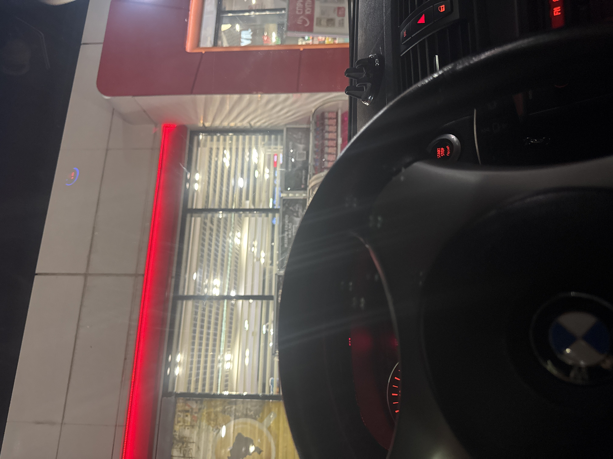 The image shows the interior of a BMW car, specifically the steering wheel with the BMW logo, the dashboard with a start/stop engine button, and the driver's view through the windshield. The location appears to be in front of a building with a red facade and large vertical windows, likely a shop or commercial establishment. The scene suggests the car is parked in front of the building.