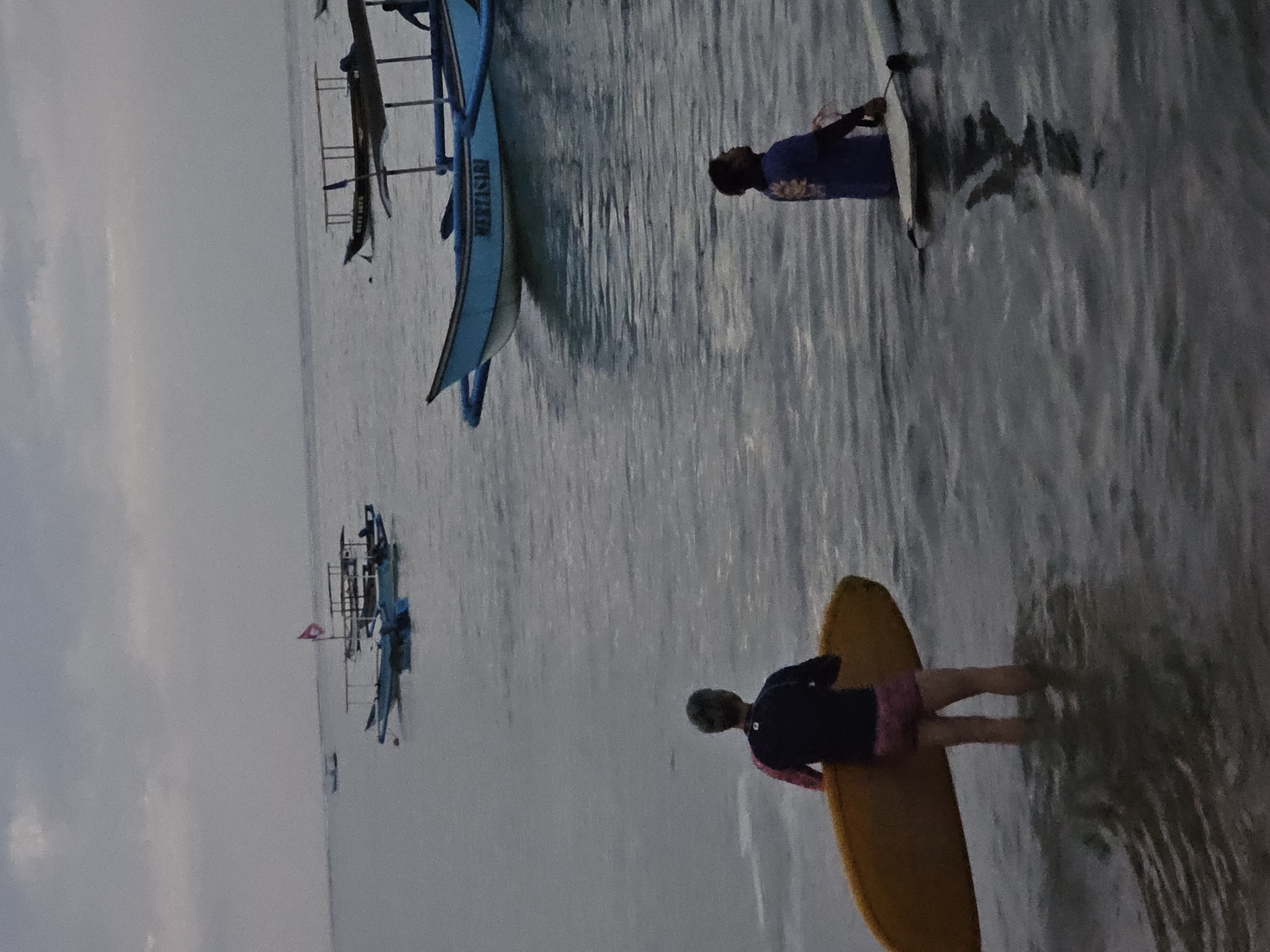In Denpasar, Indonesia, at dusk, two individuals are in the shallow ocean near a beach, each with a surfboard. The person on the left, facing away from the camera, is a surfer with graying hair, wearing a dark long-sleeved rashguard with pink accents and pink shorts. They are holding a yellow surfboard vertically. The person on the right, also facing away, is wearing a blue short-sleeved shirt and dark shorts, and is standing near a white surfboard in the water. Several traditional Indonesian fishing boats are moored in the background, some with canopies. The sky is overcast, and the water is calm.